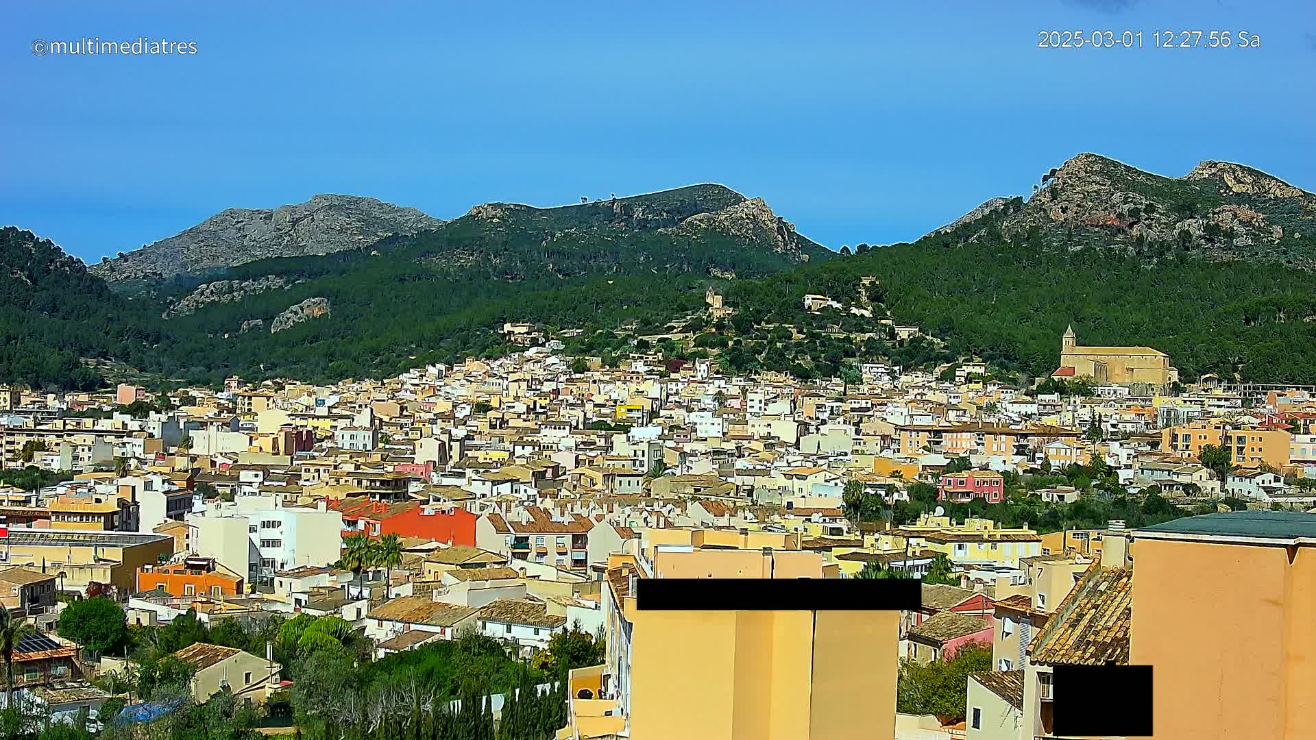 A sprawling town of low-rise buildings nestles at the base of verdant, rocky mountains under a clear blue sky.