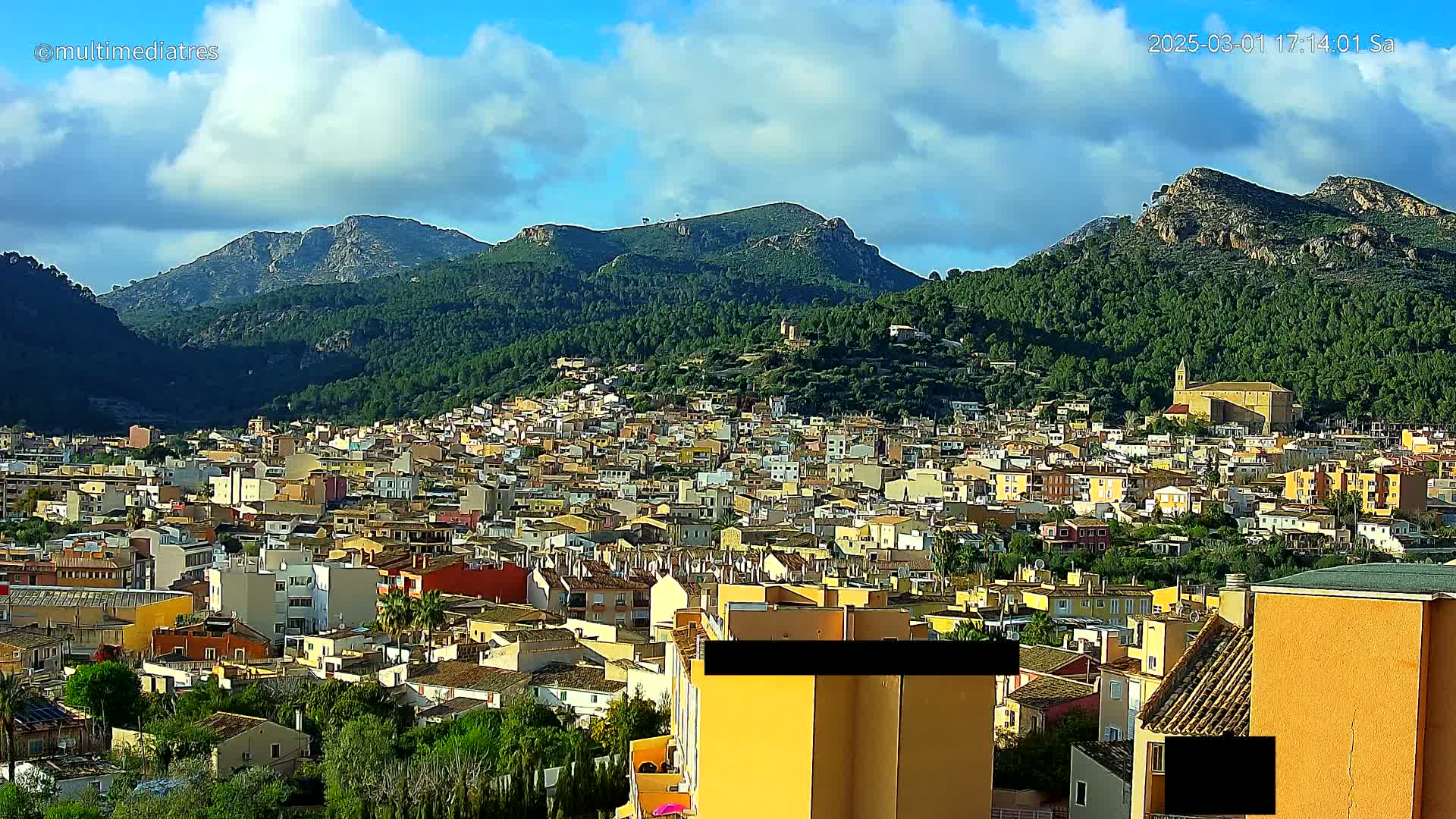 A partly cloudy day reveals a sprawling town nestled in a valley, surrounded by lush green hills and rocky mountains.