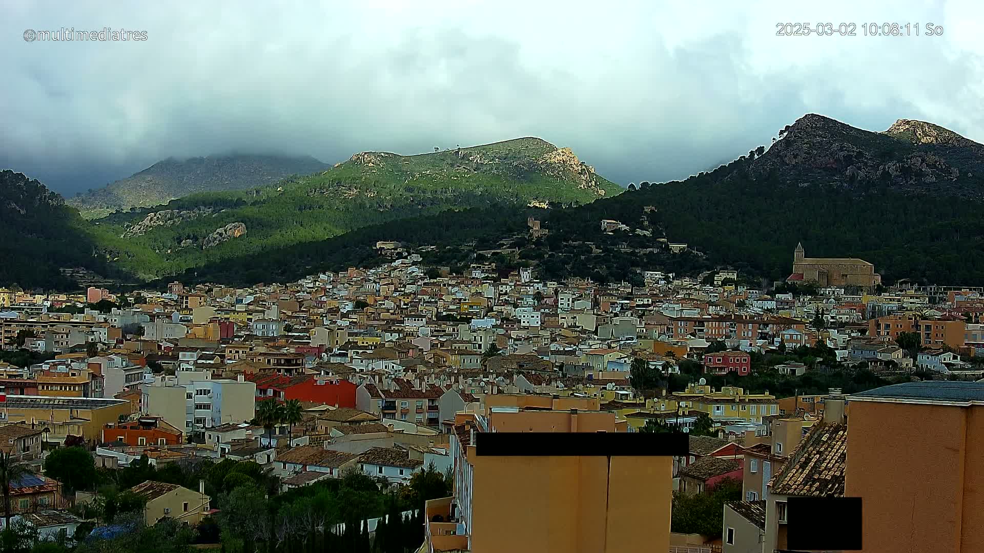 A sprawling town of low-rise buildings nestles in a valley between verdant, mountainous terrain under a mostly cloudy sky.