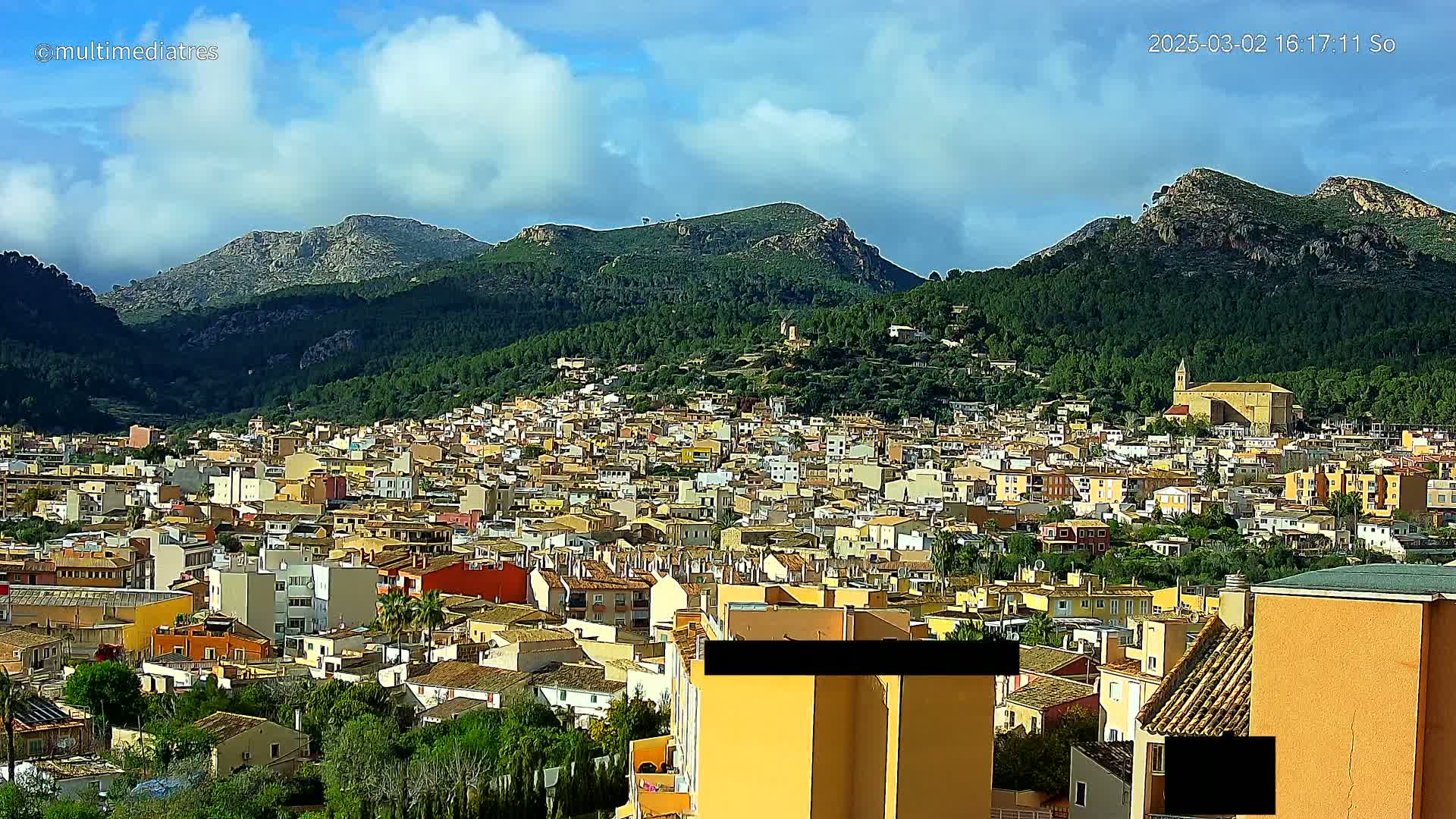 A sprawling town of low-rise buildings nestled in a valley between verdant, mountainous terrain under a partly cloudy sky.