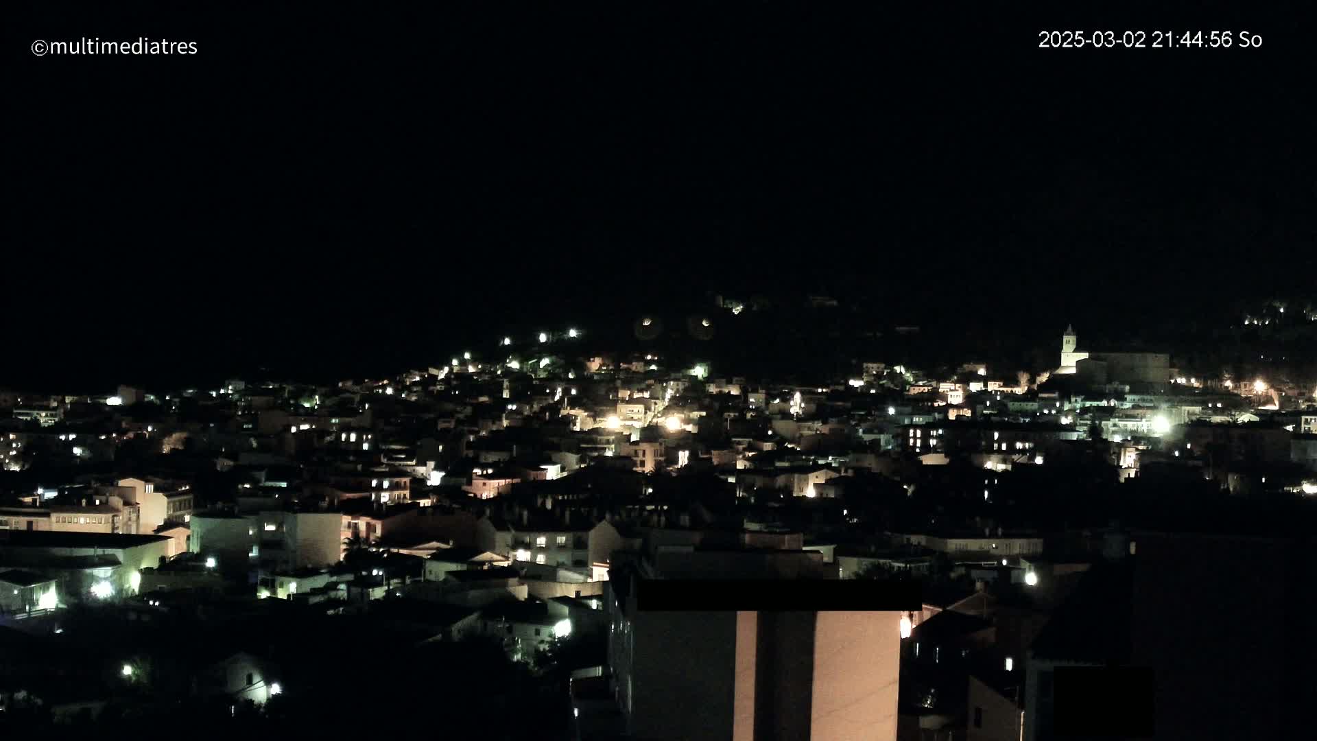 A nighttime, aerial view of a town nestled on a hillside, illuminated by scattered lights.