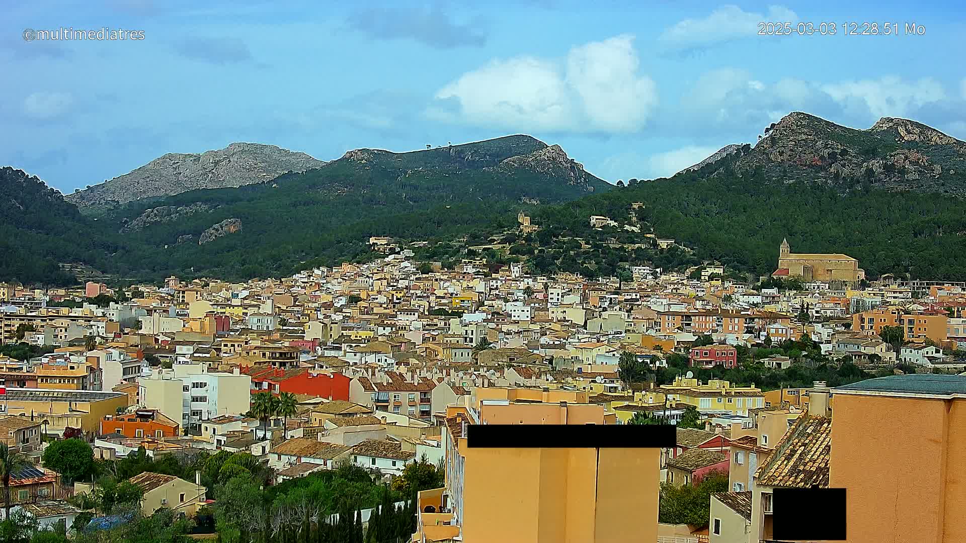 A sprawling town of tan and orange buildings nestled in a valley between lush green mountains under a mostly sunny sky.