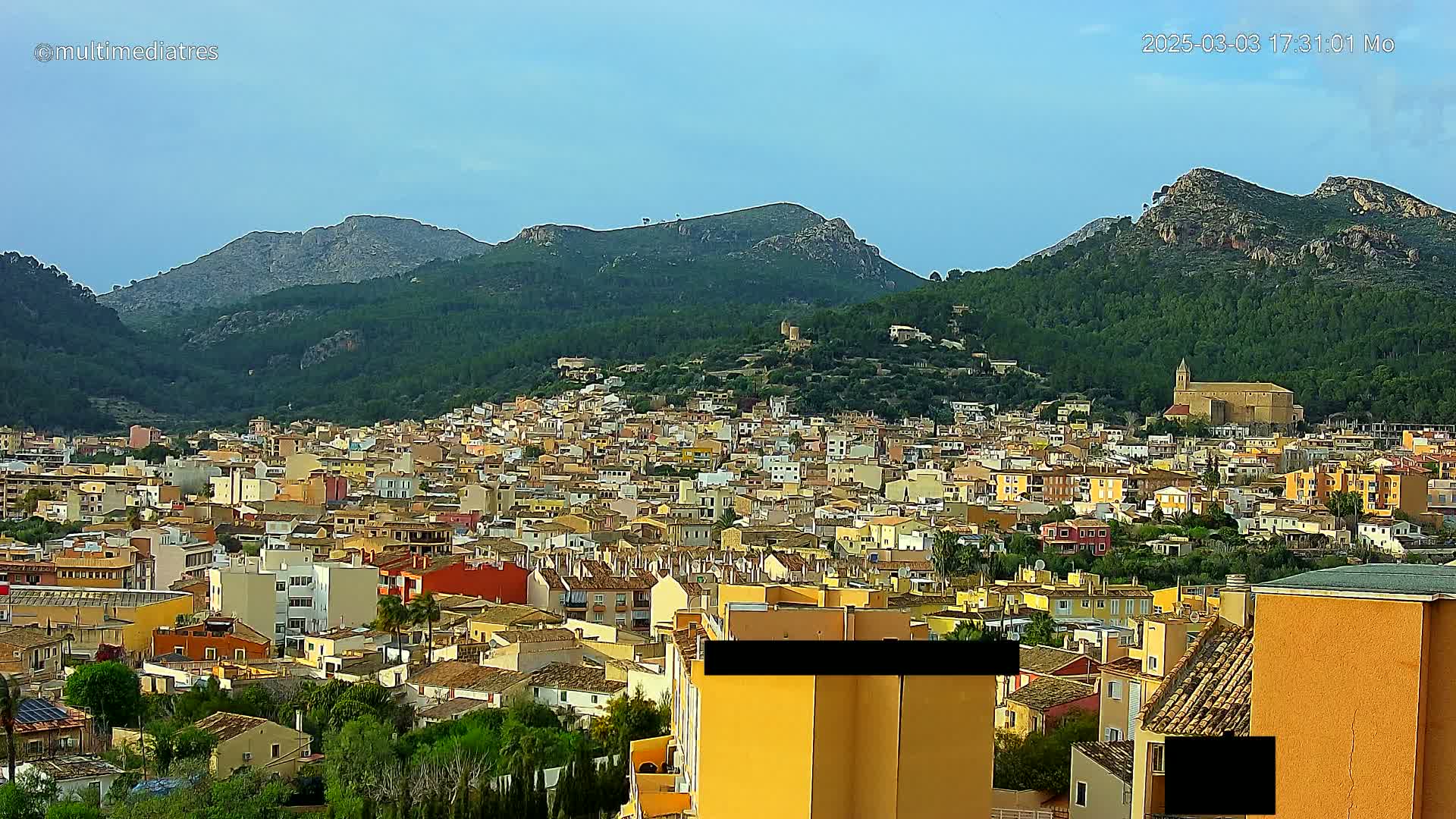 A sprawling town of low-rise buildings nestles in a valley surrounded by green, mountainous terrain under a mostly clear sky.