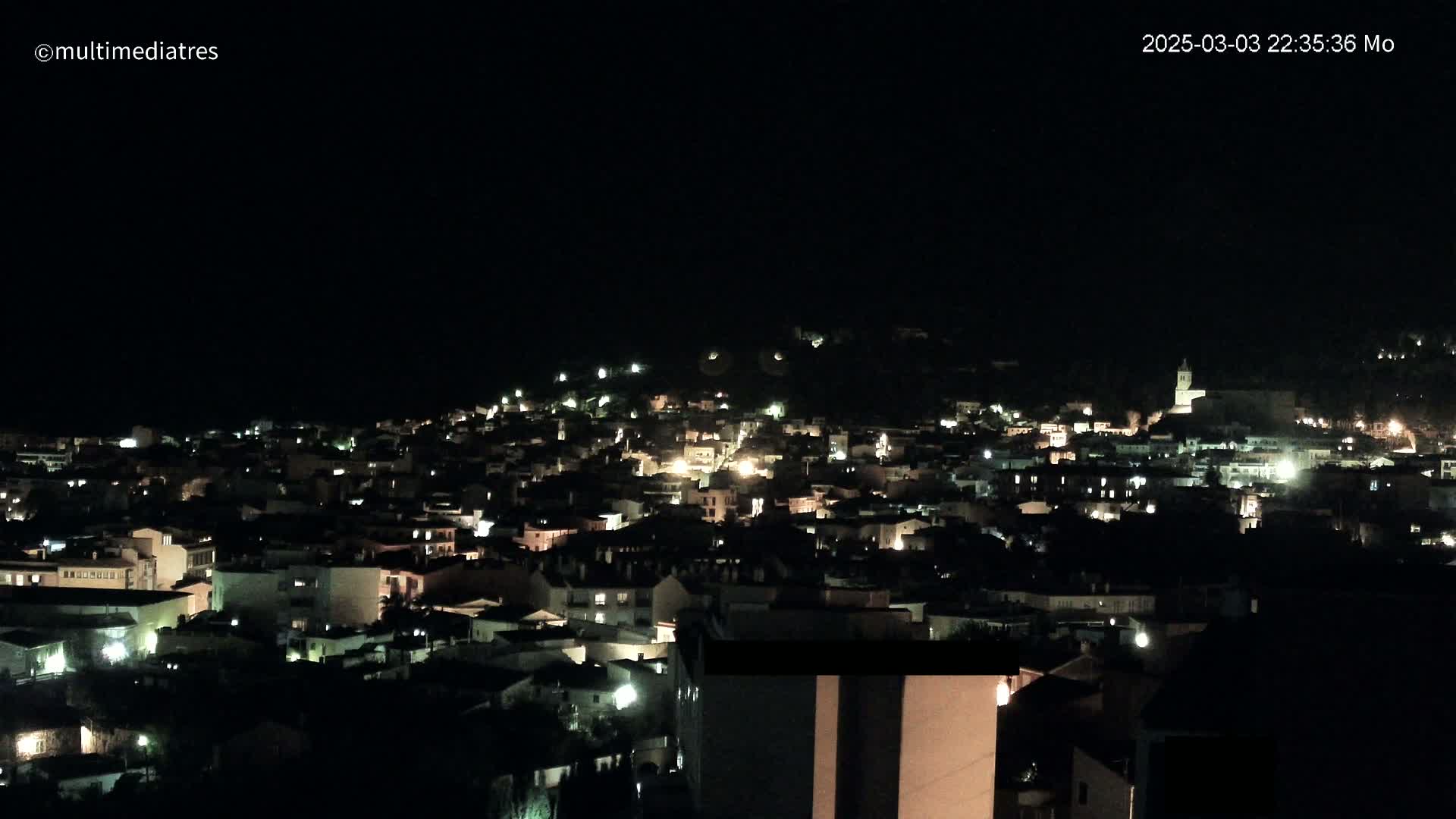 A nighttime, aerial view shows a town nestled on a hillside, with many lights illuminating buildings and streets under a clear, dark sky.