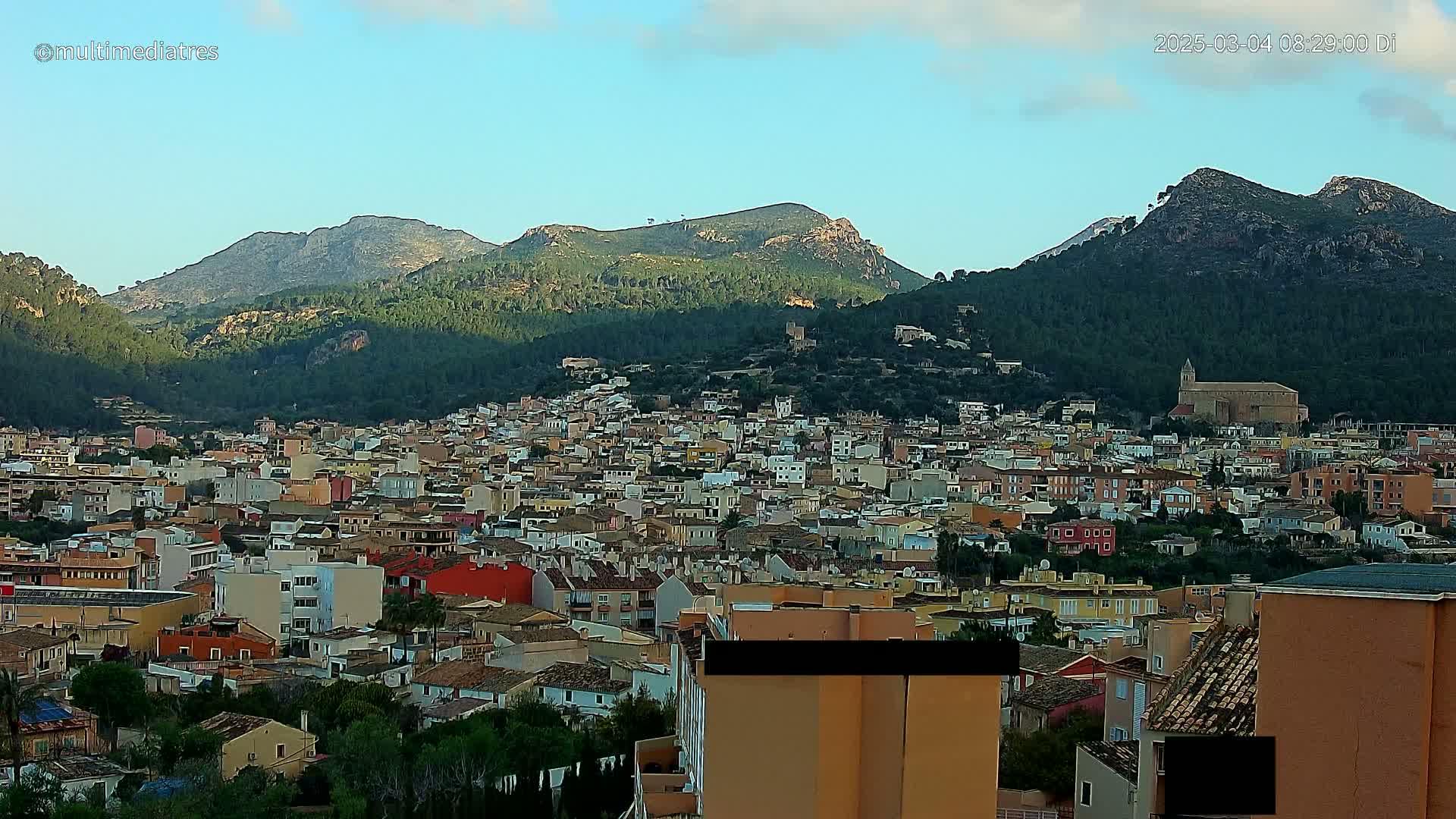 A sprawling town of low-rise buildings nestles at the foot of verdant, rocky mountains under a mostly clear, light-blue sky.