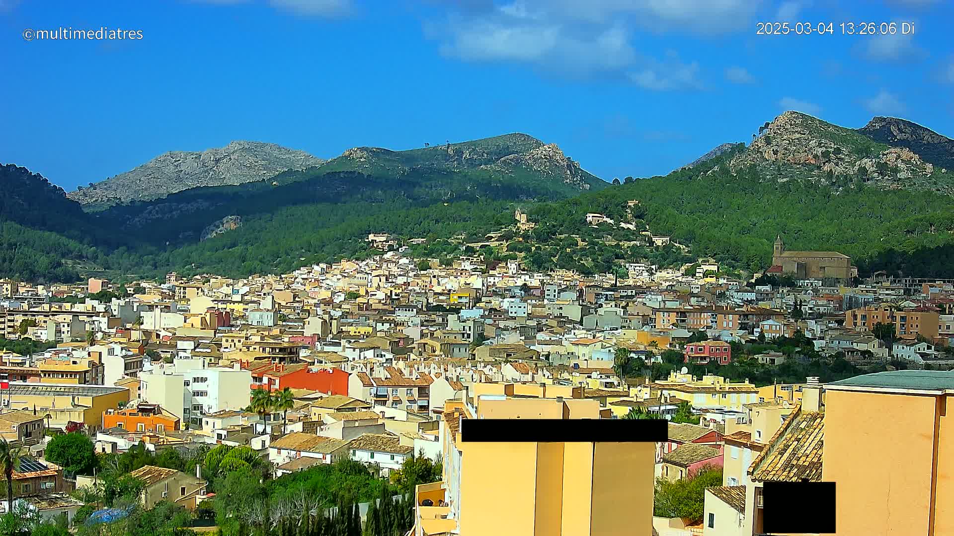 A sprawling town of low-rise buildings nestled against a backdrop of verdant, mountainous terrain under a bright blue sky with a few scattered clouds.