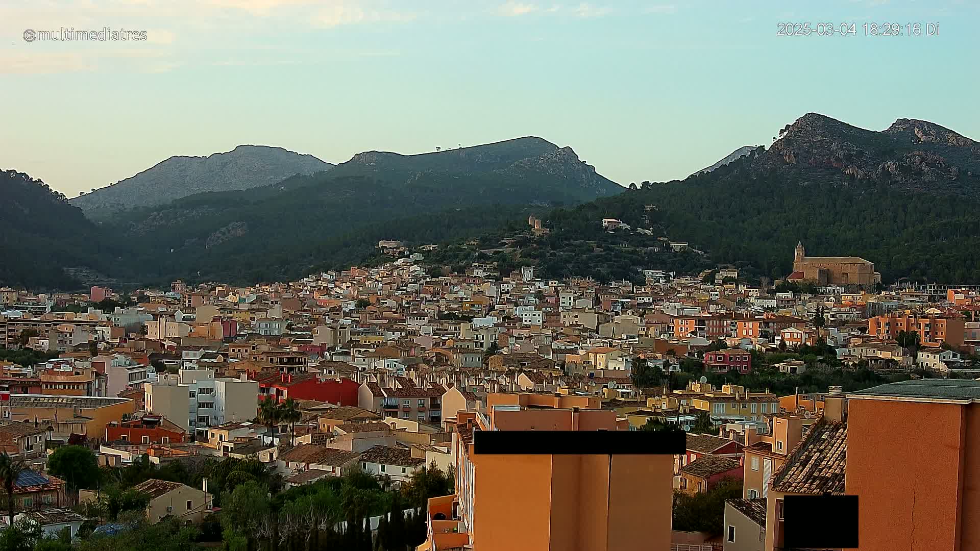 A dense collection of low-rise buildings in a town nestled against a backdrop of mountainous terrain under a clear, light-blue sky.