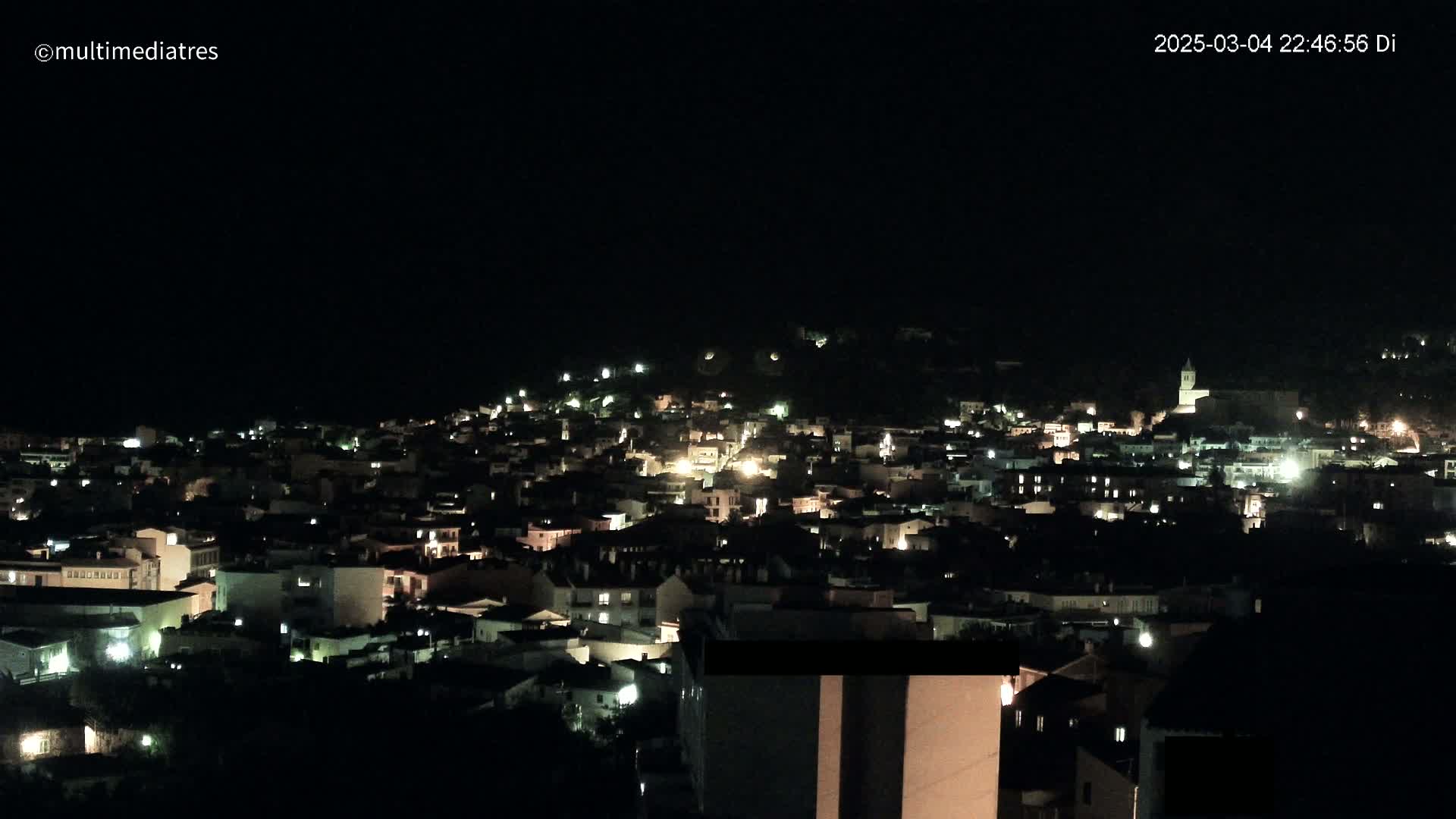 A nighttime view of a town nestled on a hill, illuminated by scattered lights under a dark, clear sky.