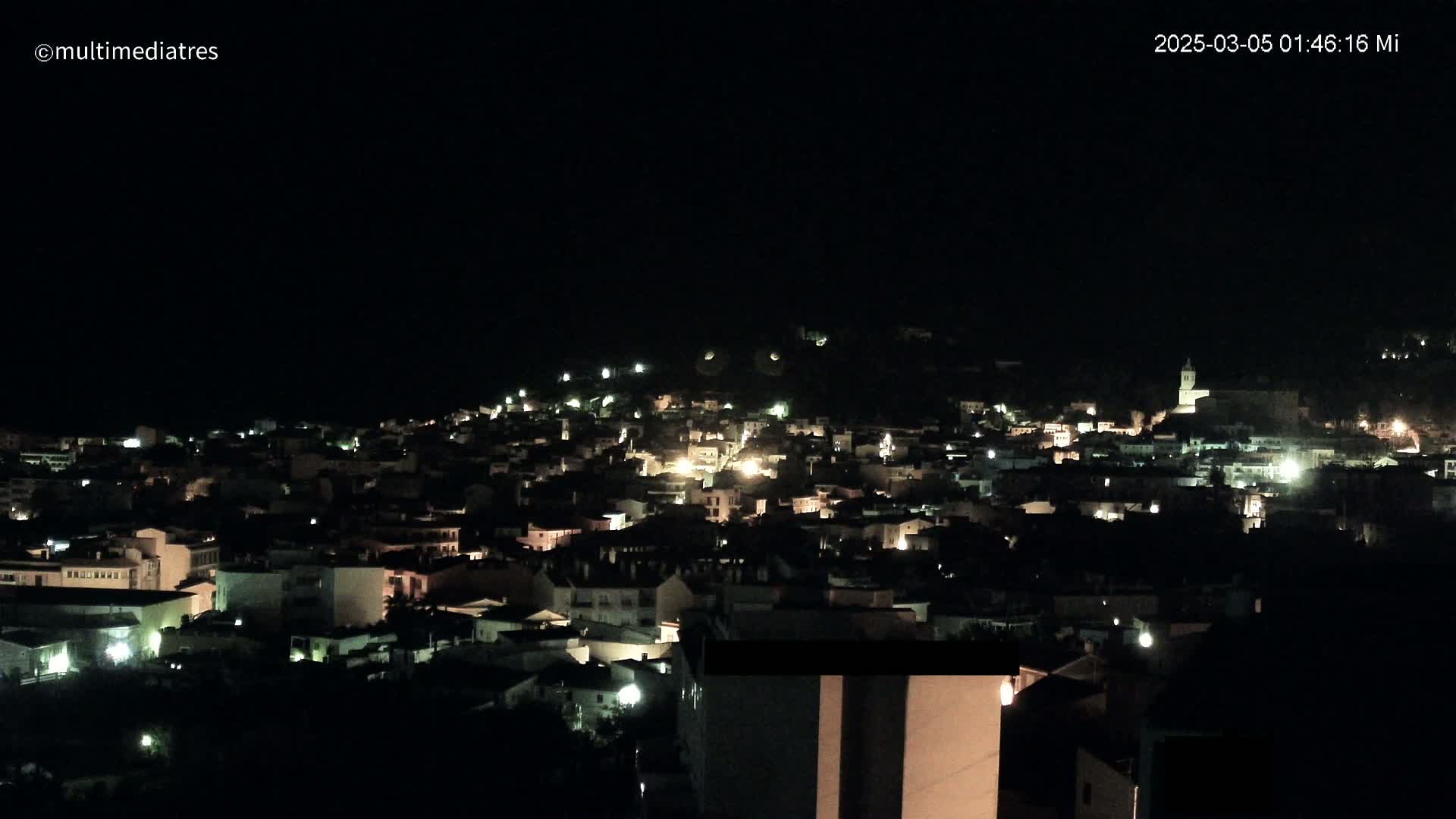 A nighttime view of a town nestled on a hillside, illuminated by scattered lights under a clear, dark sky.