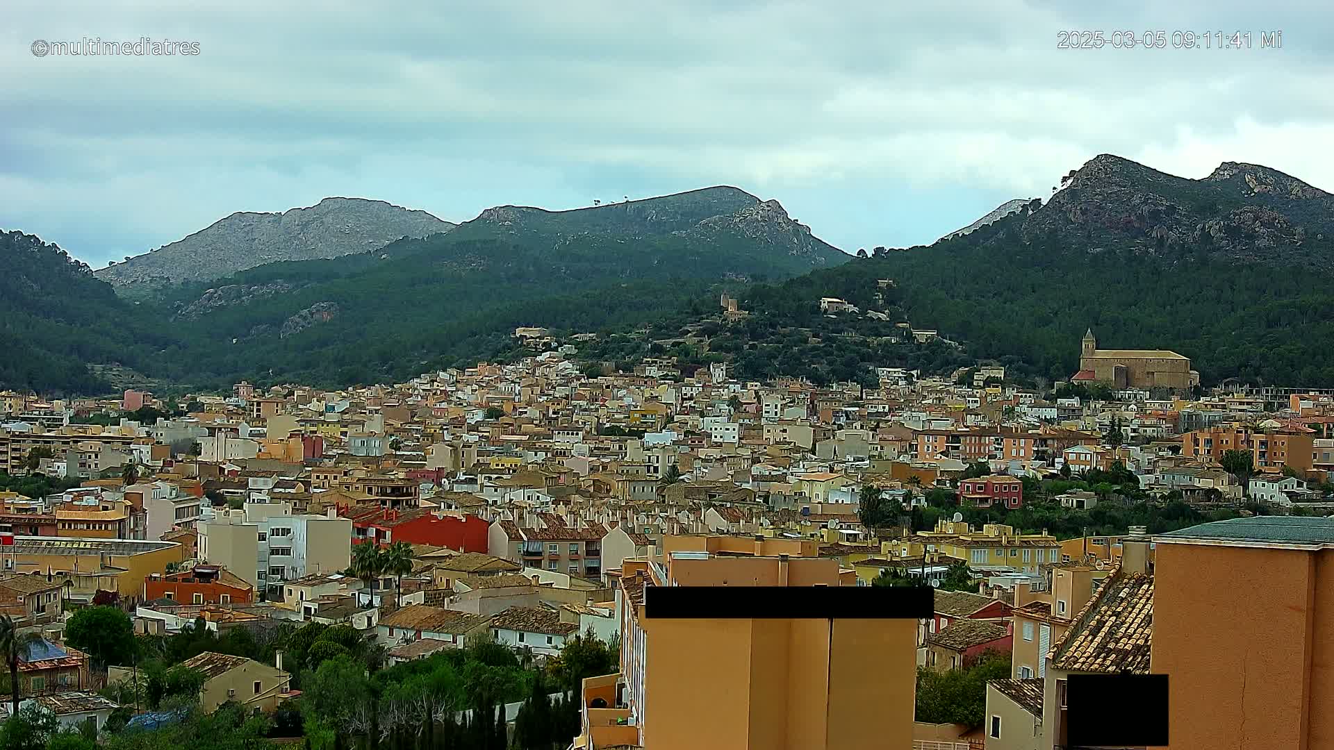 A sprawling town of tan and orange buildings nestles in a valley surrounded by verdant, mountainous terrain under a cloudy sky.
