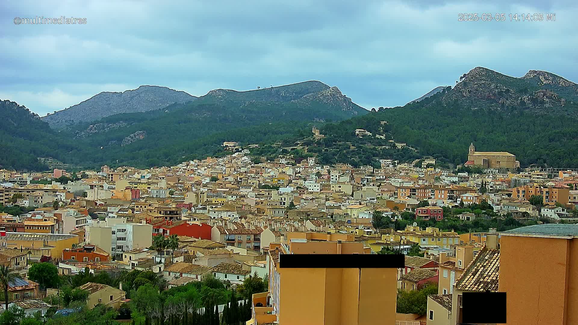 A sprawling town of low-rise buildings, nestled amongst green hills and under a mostly cloudy sky.