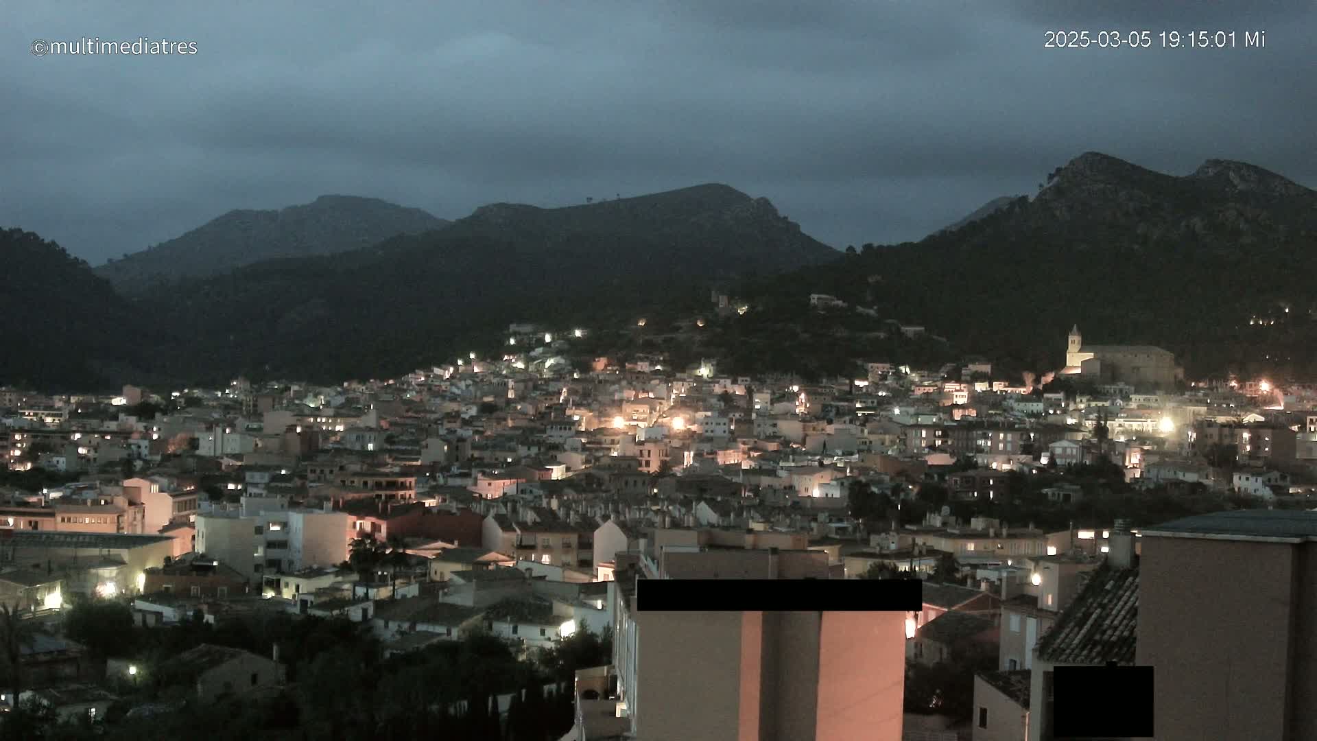 A nighttime view of a town nestled in a valley surrounded by dark mountains under a cloudy sky.