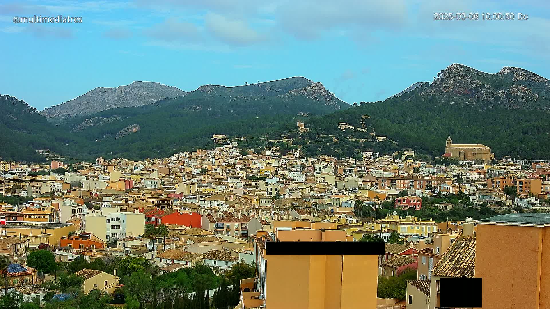 A sprawling town of low-rise buildings, nestled in a valley between verdant, mountainous terrain under a mostly clear sky.