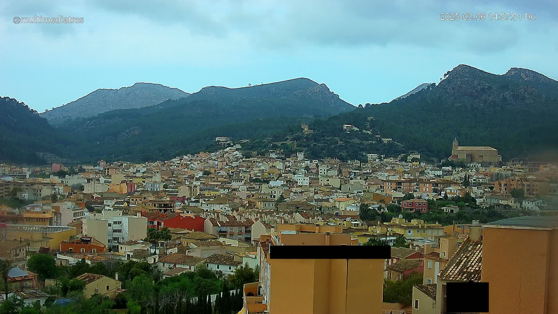 A sprawling town of tan and red-roofed buildings nestled in a valley between verdant, mountainous terrain under an overcast sky.