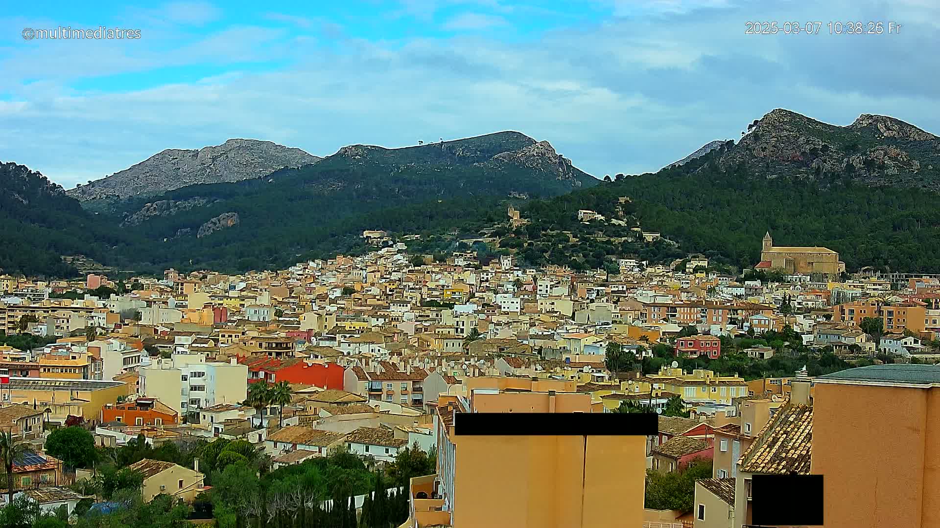 A sprawling town of low-rise buildings in warm colors nestles against the foothills of a mountain range under a partly cloudy sky.