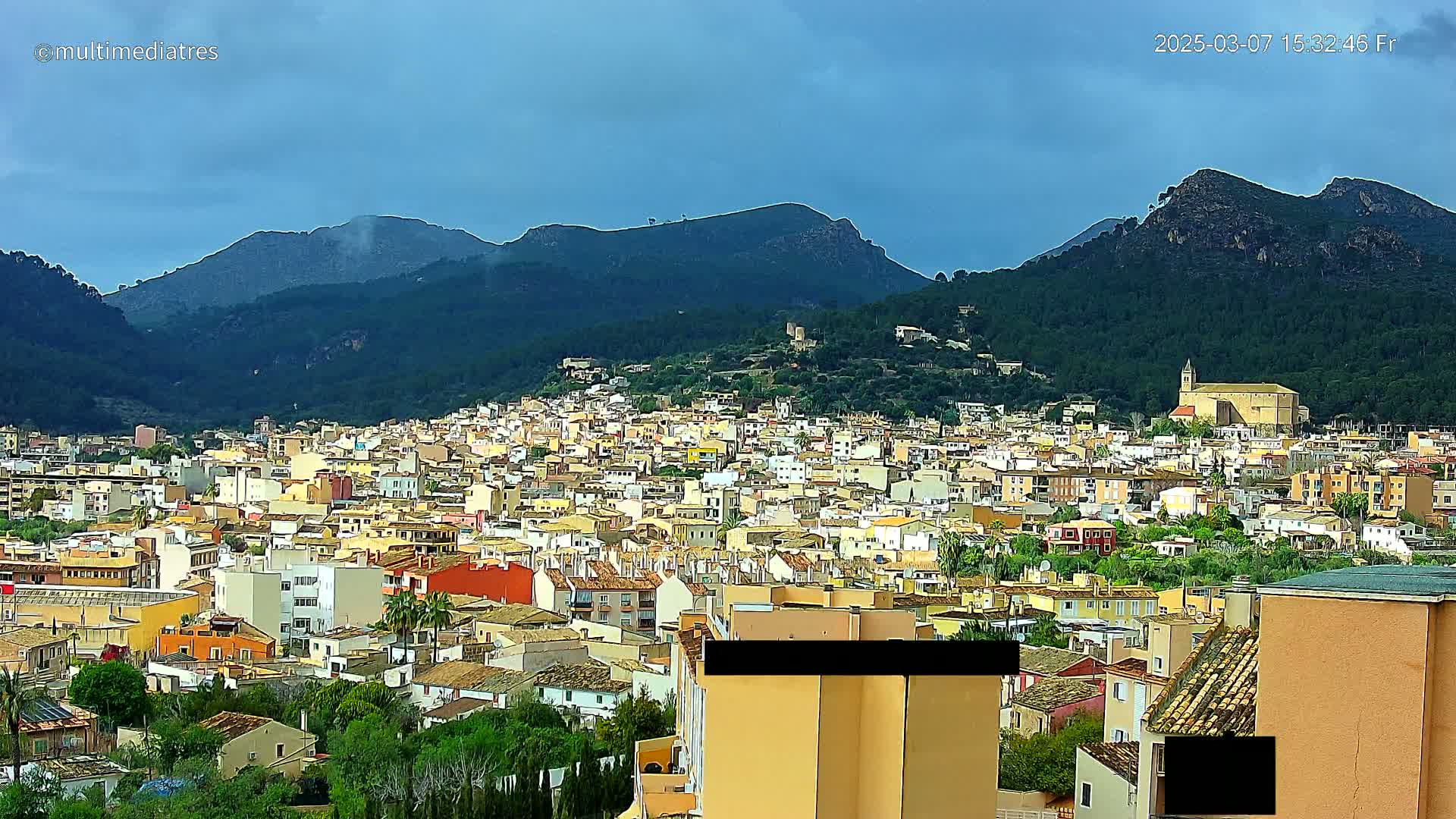 A sprawling town of low-rise buildings nestled in a valley, surrounded by lush green mountains under a partly cloudy sky.