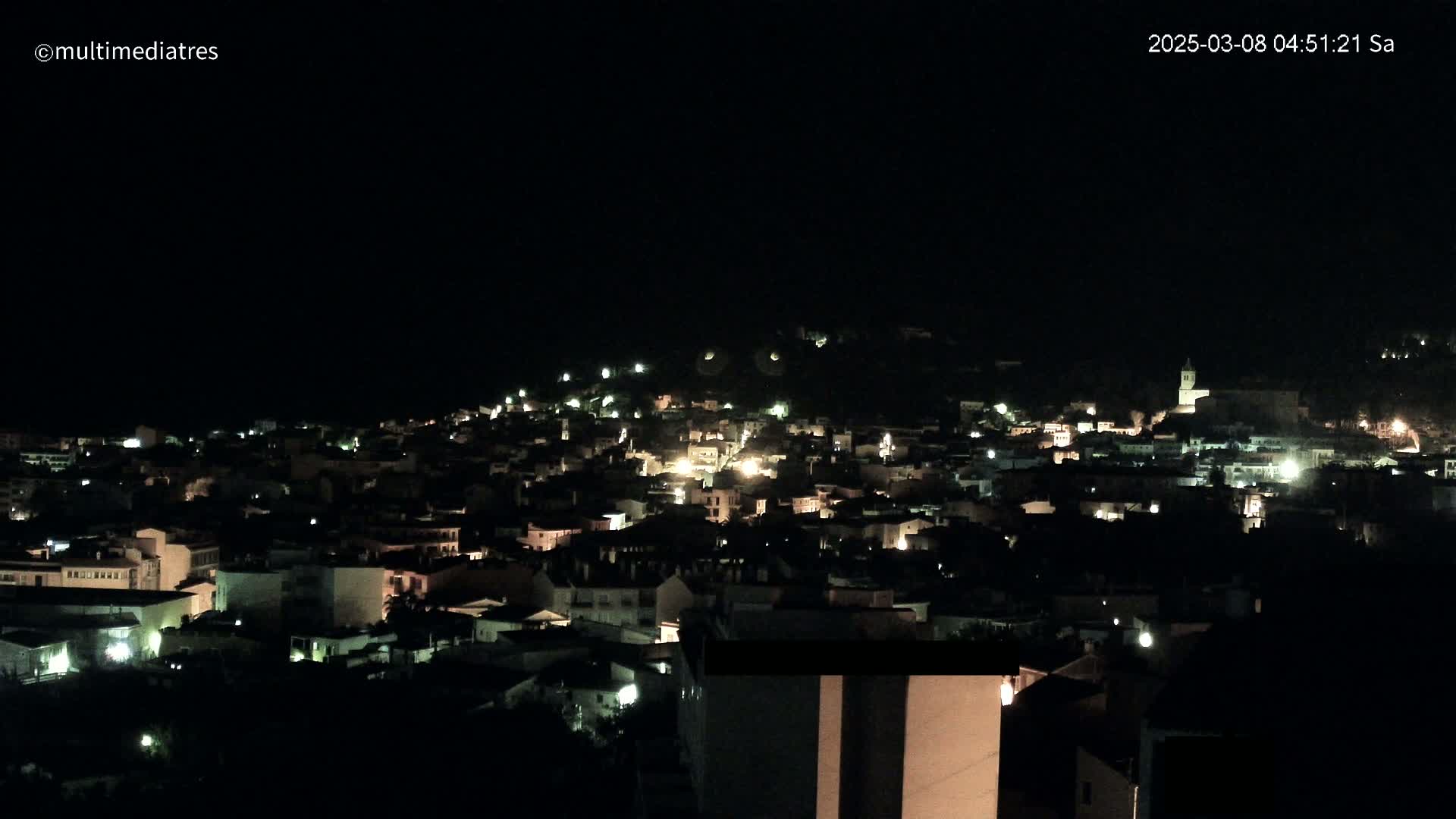 A nighttime, aerial view of a town nestled on a hill, with scattered lights illuminating the buildings.