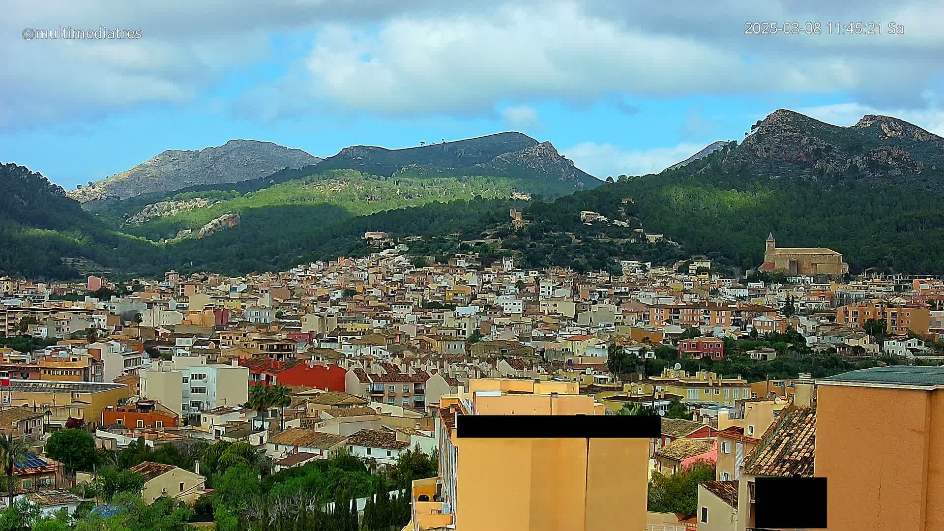 A sprawling town of numerous tan and red-roofed buildings nestles in a valley surrounded by verdant, rocky mountains under a partly cloudy sky.