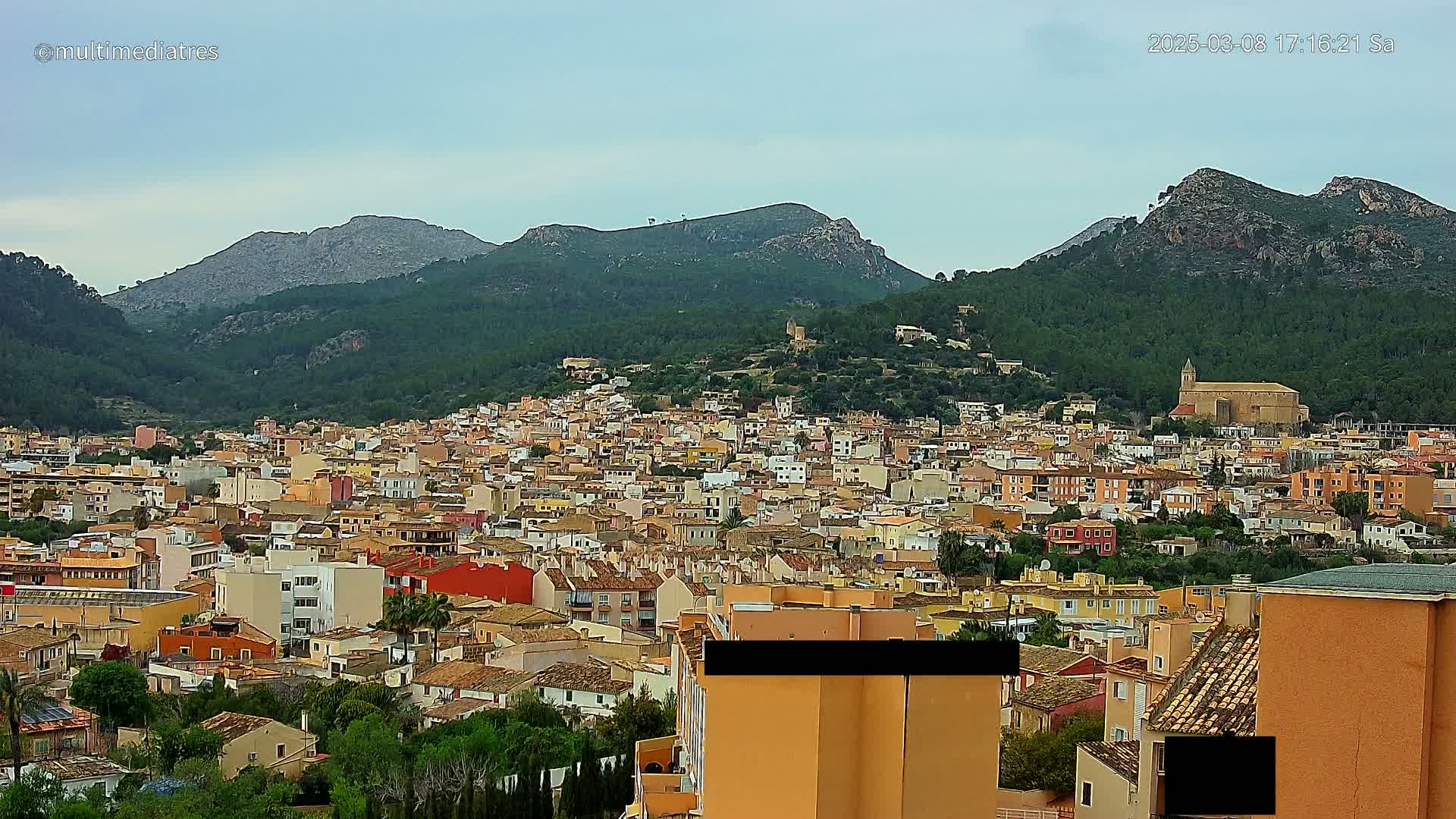 A sprawling town of low-rise buildings climbs a hillside beneath a mostly cloudy sky, backed by several verdant mountains.
