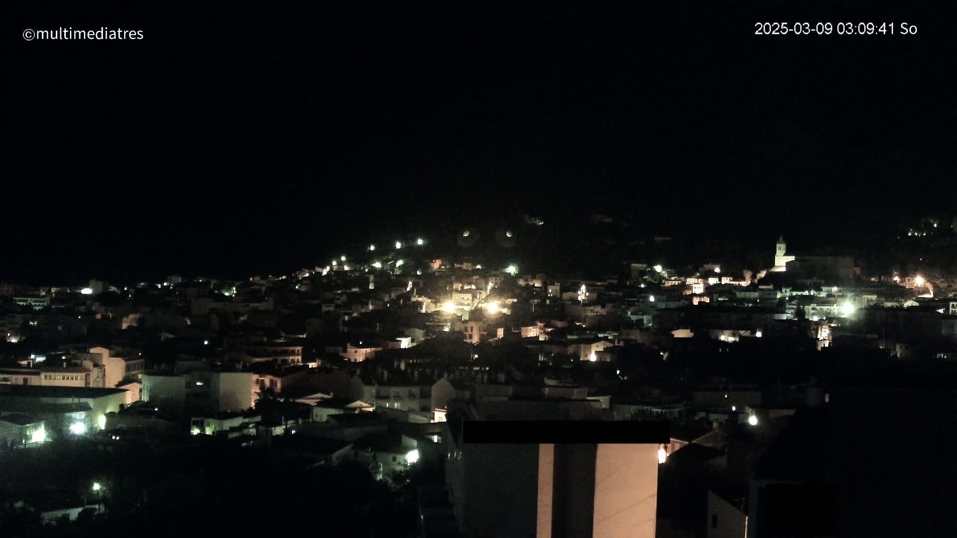 A nighttime view of a town nestled on a hillside, illuminated by scattered lights under a dark sky.