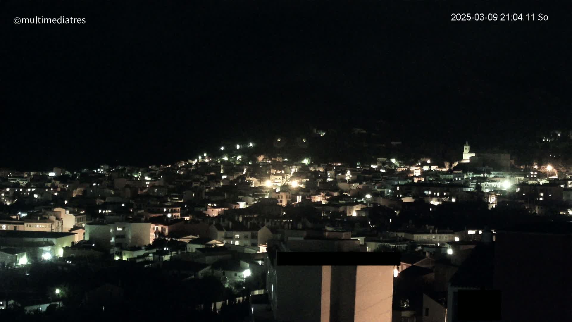 A nighttime view of a town illuminated by scattered lights on a hill under a clear, dark sky.