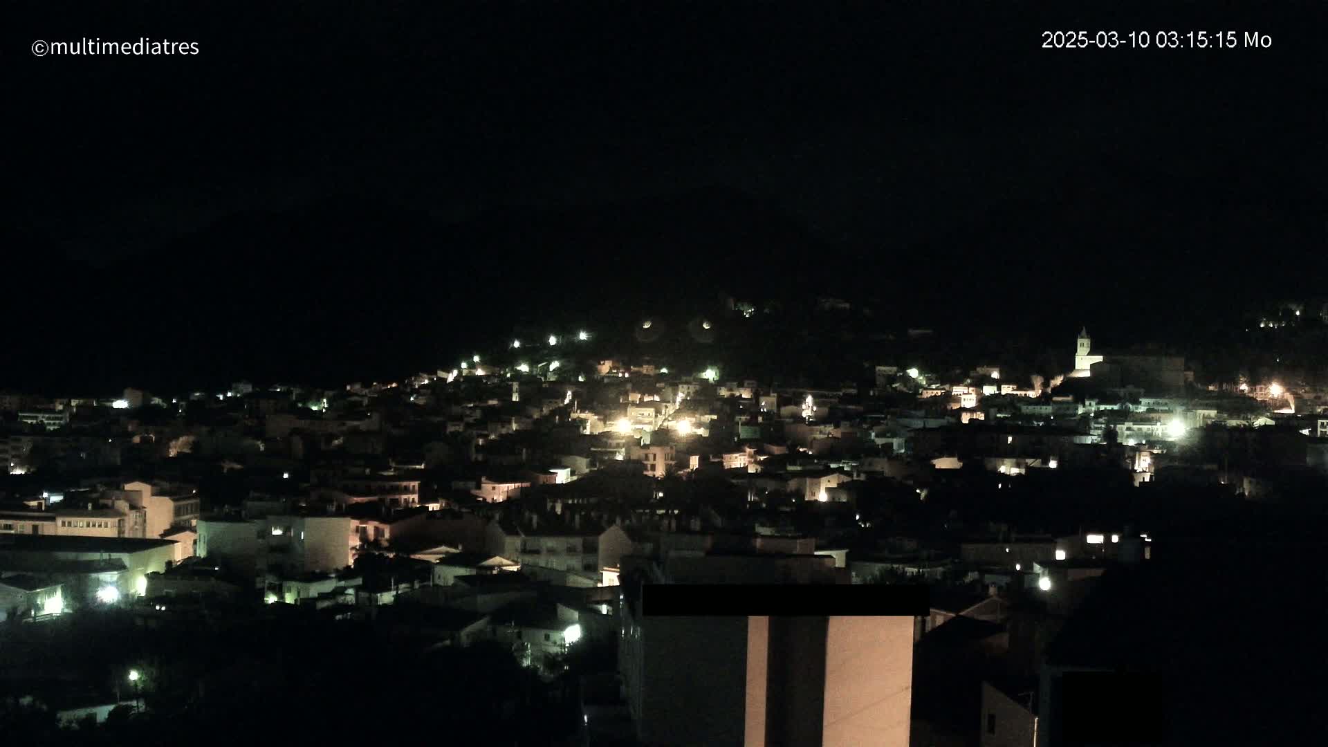 A nighttime view of a town nestled on a hill, illuminated by scattered lights under a dark, clear sky.