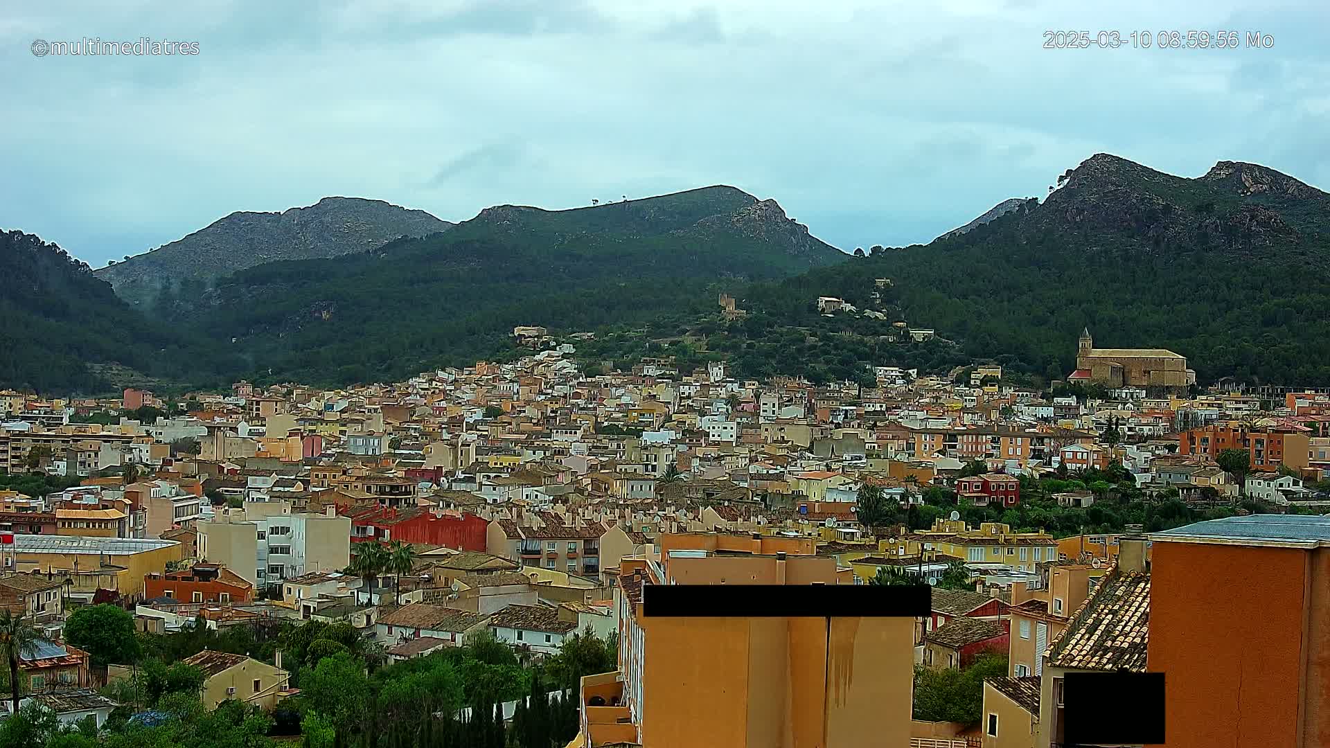 A sprawling town of colorful buildings nestles in a valley surrounded by lush green mountains under a cloudy sky.