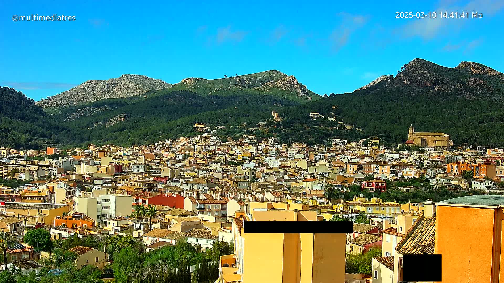 A sprawling town of tan and orange buildings nestles at the base of a verdant mountain range under a clear blue sky.