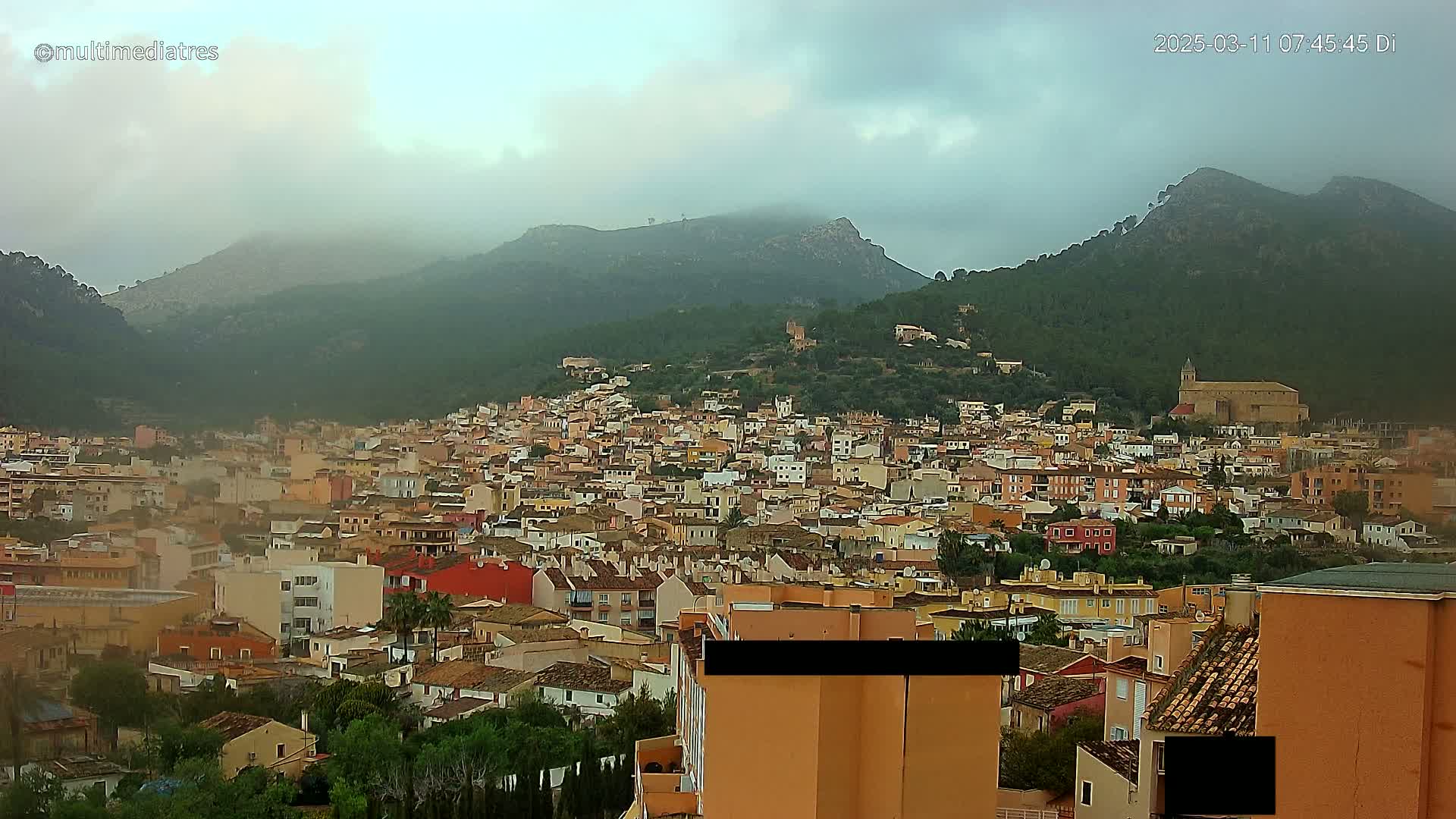 A dense, hilly town of low-rise buildings, nestled against mist-covered mountains under an overcast sky.