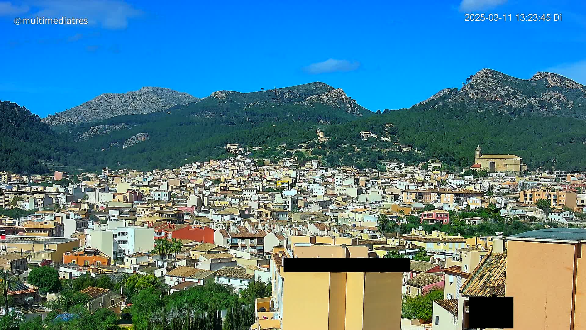 A sprawling town nestles at the base of verdant, rocky mountains under a clear blue sky.
