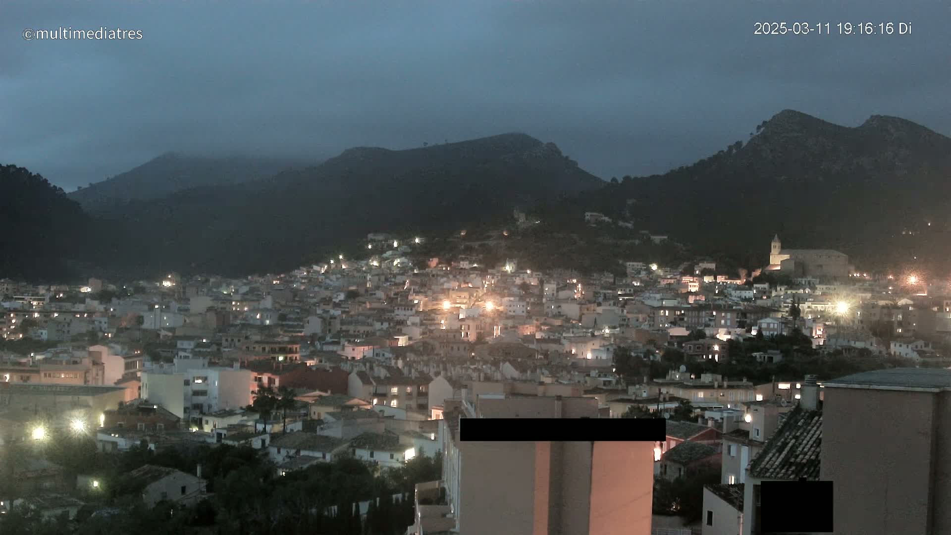A nighttime view of a town nestled in a valley between dark, mountainous terrain under a cloudy, dusky sky.
