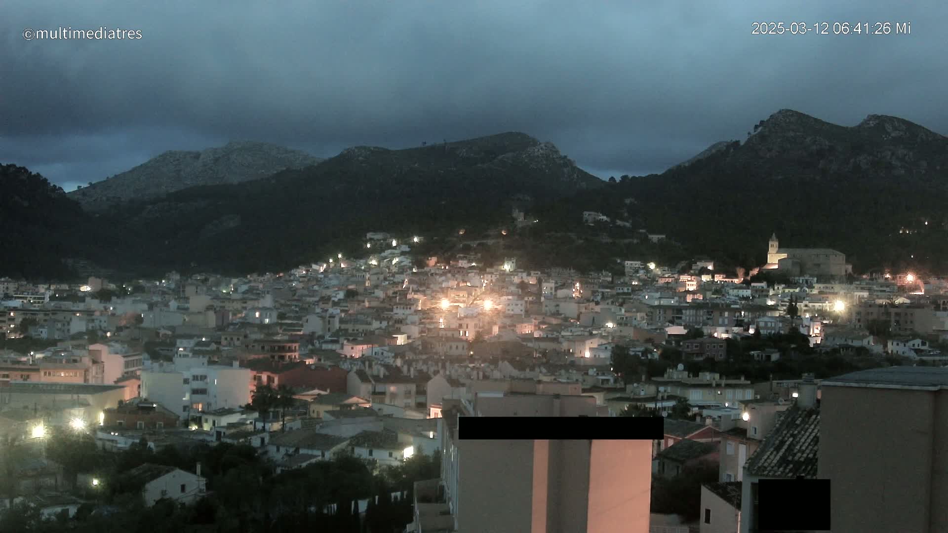 A nighttime view of a town nestled in a mountain valley under a dark, cloudy sky.