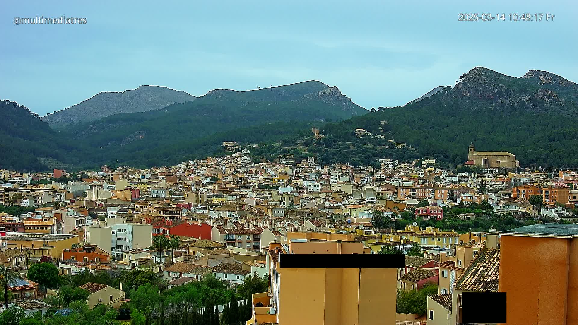 A sprawling town of multi-colored buildings nestled in a valley surrounded by verdant, mountainous terrain under a mostly cloudy sky.