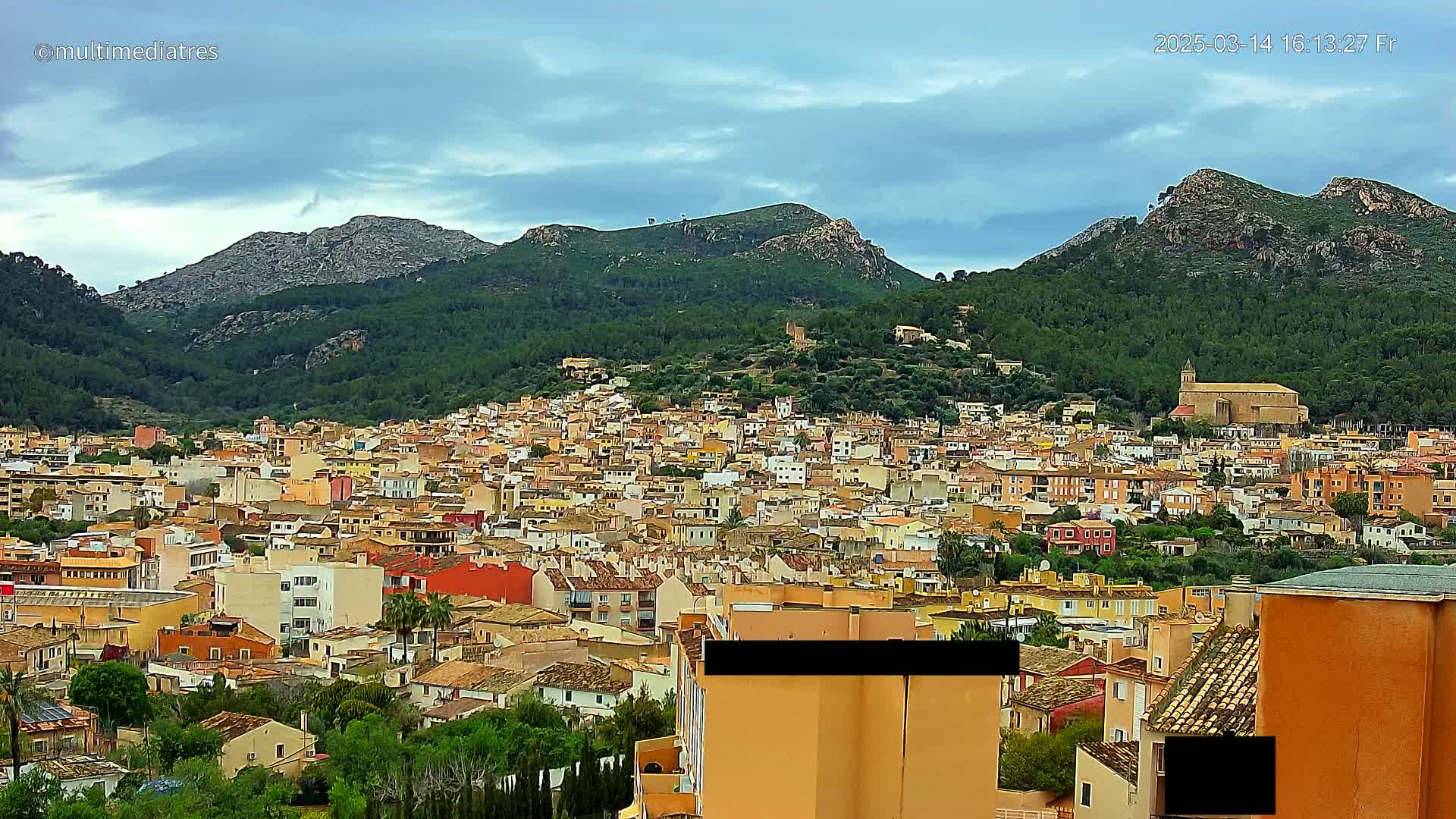 A dense collection of buildings, mostly low-rise and earth-toned, stretches across a hillside beneath mountains under a partly cloudy sky.