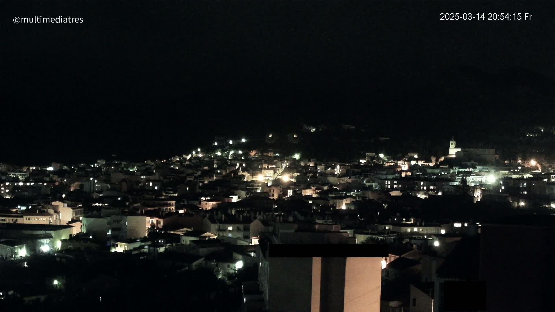 A nighttime view of a town nestled on a hillside, with numerous lights illuminating the buildings.