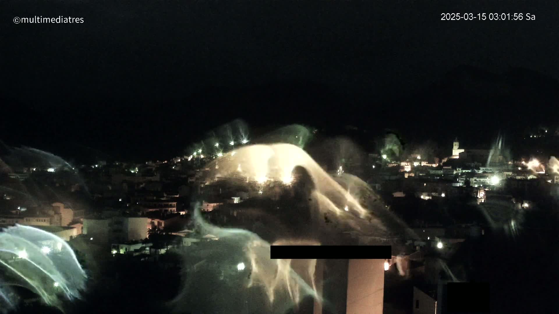 A nighttime view of a town illuminated with lights, with large water fountains or sprinklers spraying water in arcs across the scene.