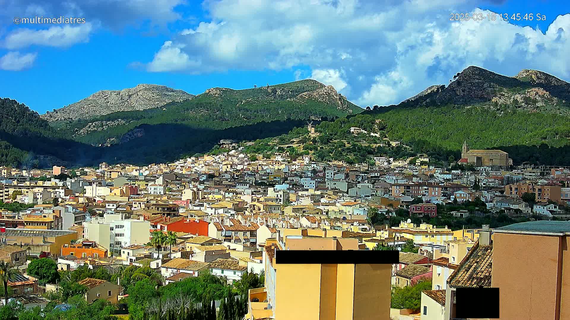 A sprawling town of low-rise buildings nestles at the base of a mountain range under a partly cloudy, sunny sky.