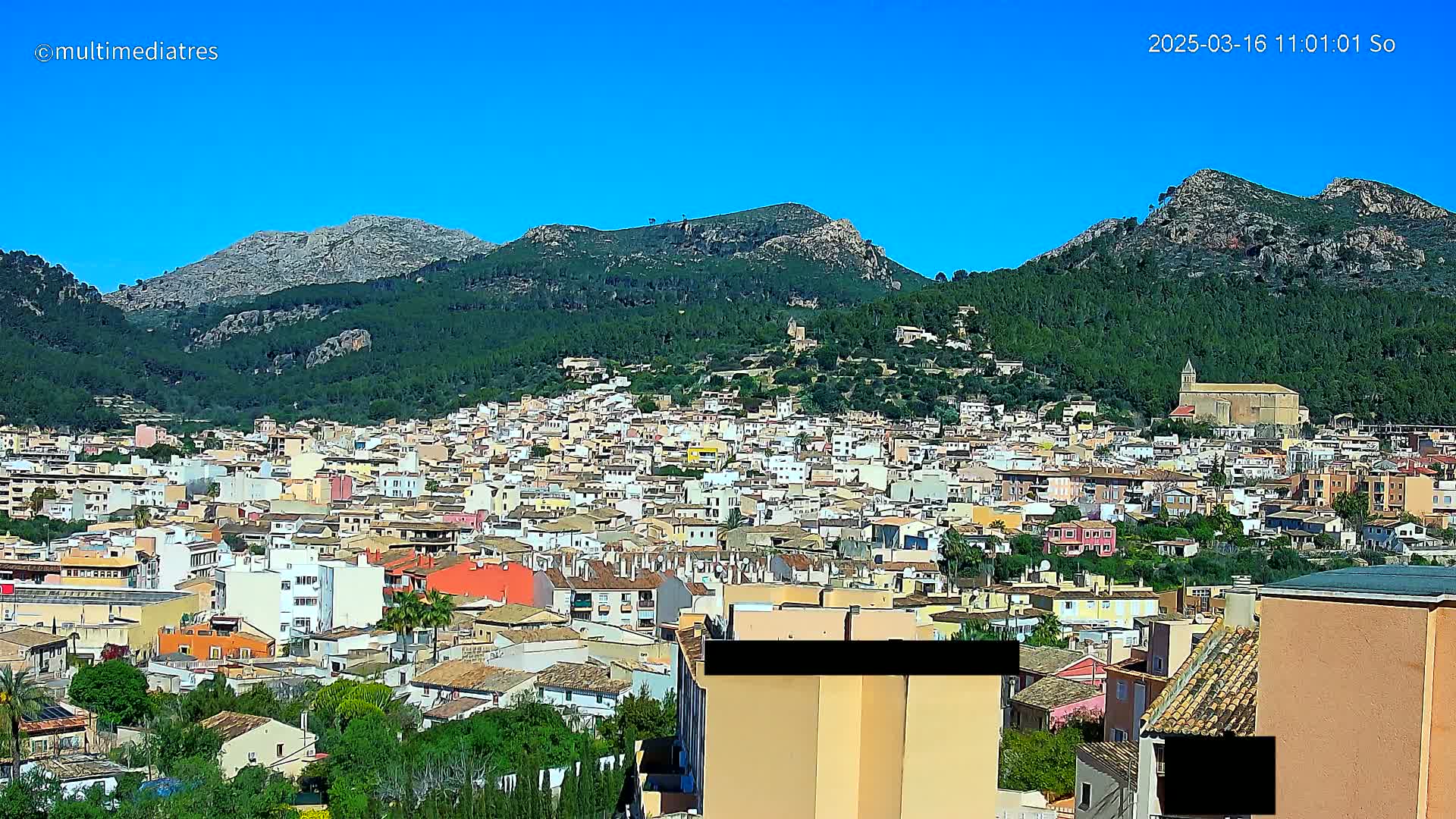 A sprawling town of closely packed buildings nestles in a valley surrounded by lush green hills and rocky mountains under a clear blue sky.