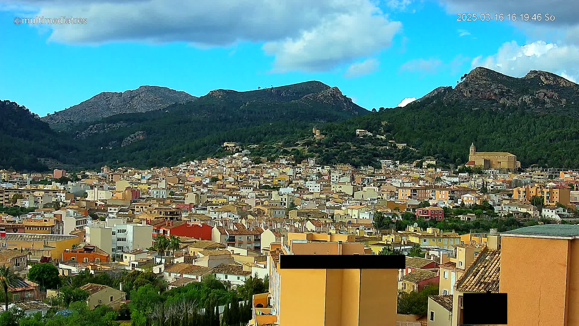 A sprawling town of low-rise buildings nestles at the foot of verdant, rocky mountains under a partly cloudy blue sky.