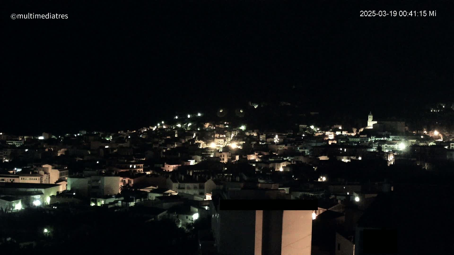 A nighttime, aerial view of a town nestled on a hillside, with scattered lights illuminating buildings.