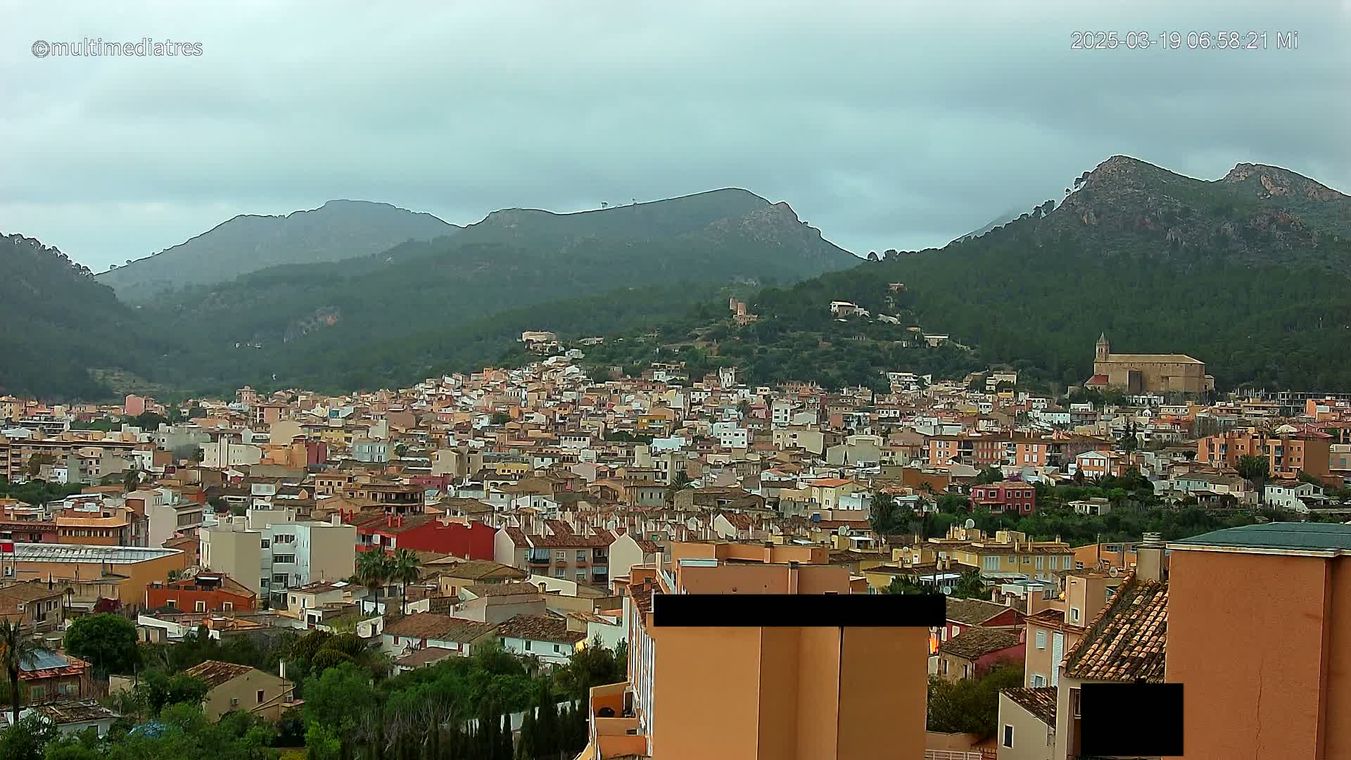 A sprawling town of tan and red buildings nestles in a valley surrounded by verdant, mountainous terrain under a cloudy sky.