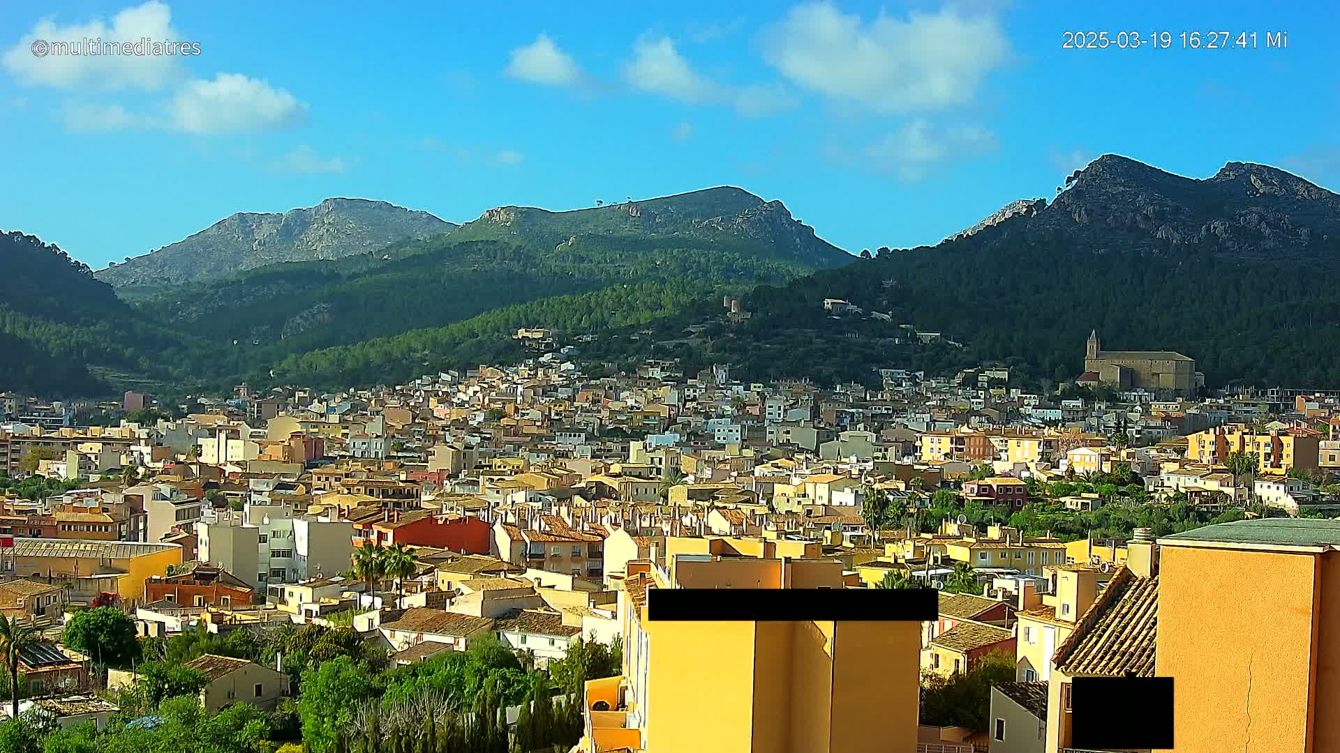 A sprawling town of tan and orange buildings nestled in a valley surrounded by lush green mountains under a mostly sunny sky.