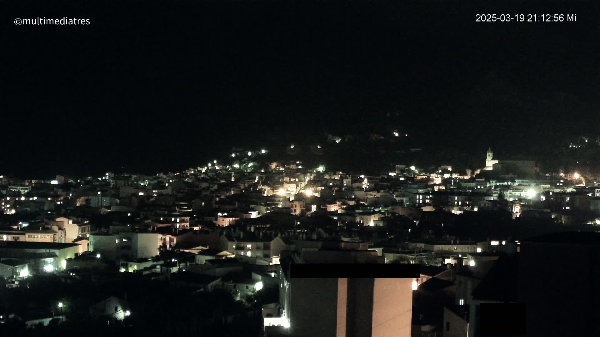 A nighttime view of a town nestled on a hill, illuminated by many small lights.