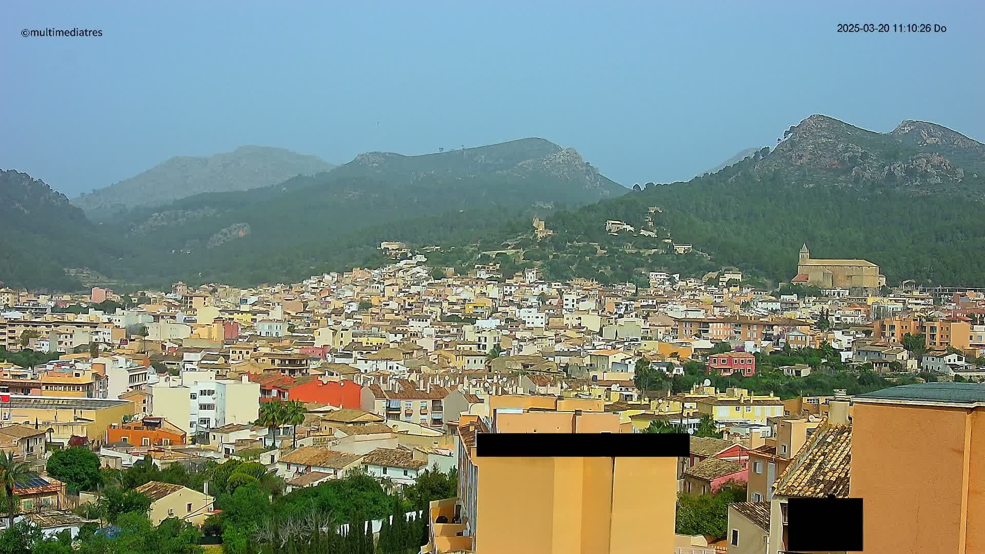 A hazy view shows a dense cluster of low-rise buildings nestled in a valley, surrounded by lush green hills and mountains under a pale sky.