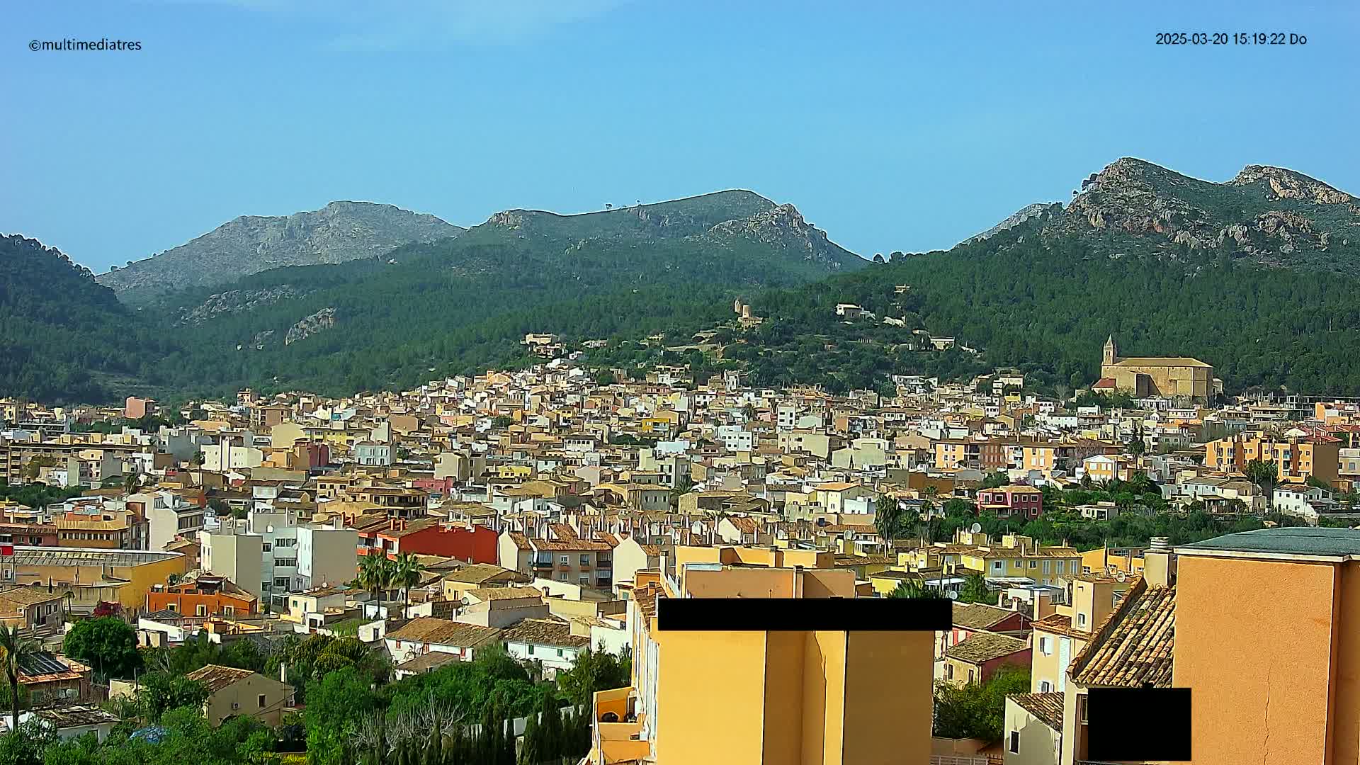 A sprawling town of low-rise buildings nestles at the foot of verdant, rocky mountains under a clear blue sky.