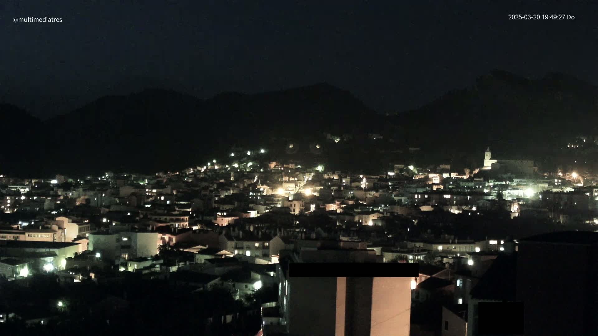 A nighttime view of a town nestled in a valley between dark mountains, illuminated by scattered lights.