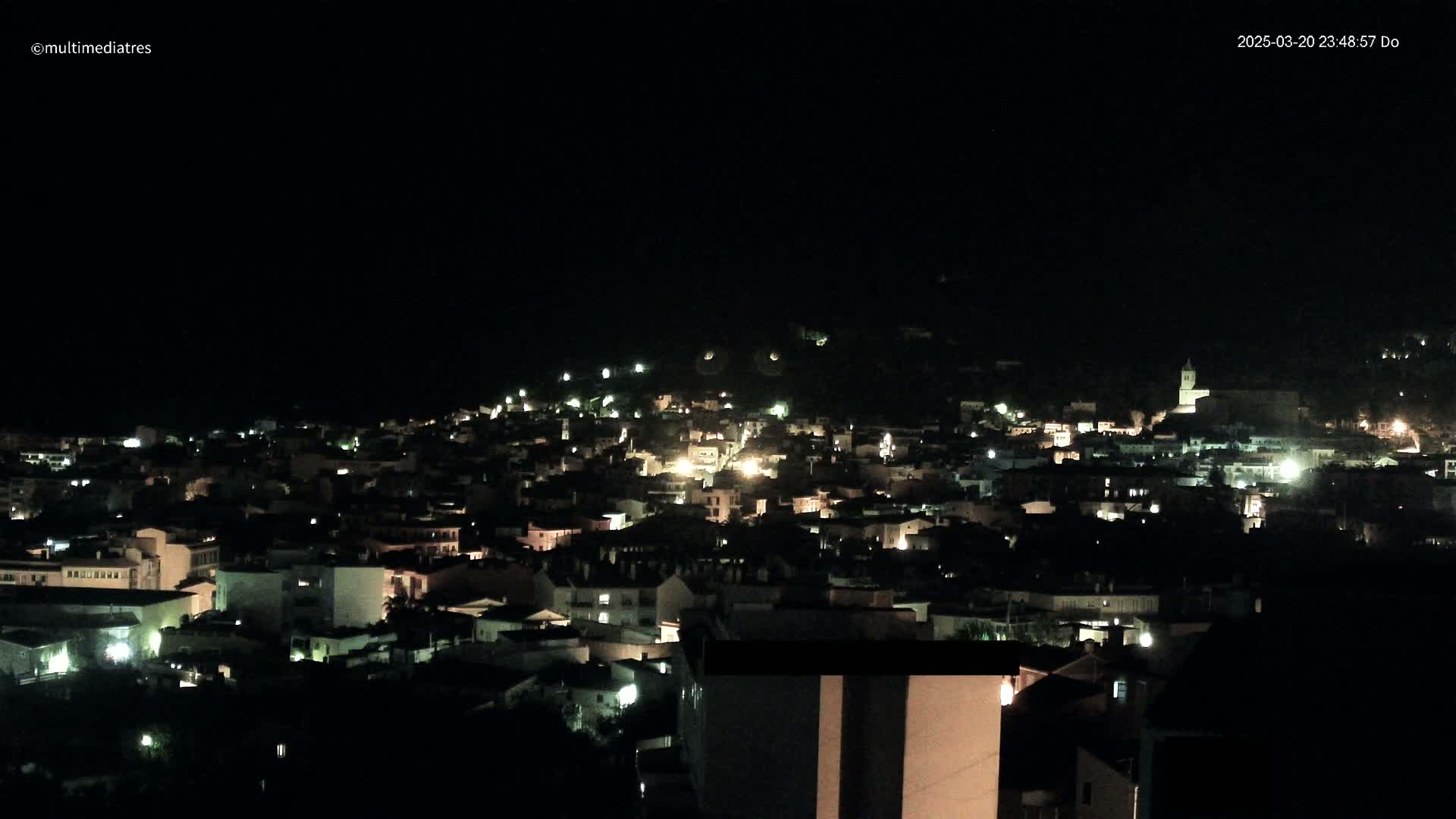 A nighttime view of a town nestled on a hillside, with scattered lights illuminating buildings under a clear, dark sky.