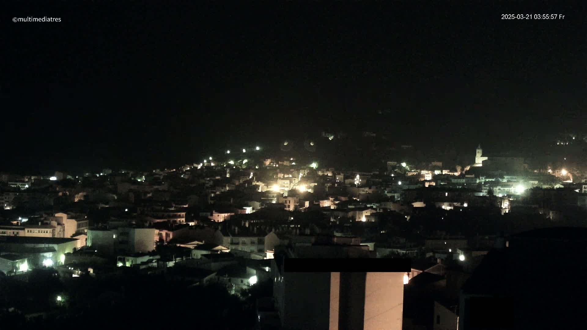 A nighttime view of a town nestled on a hillside, with numerous lights illuminating the buildings and streets.