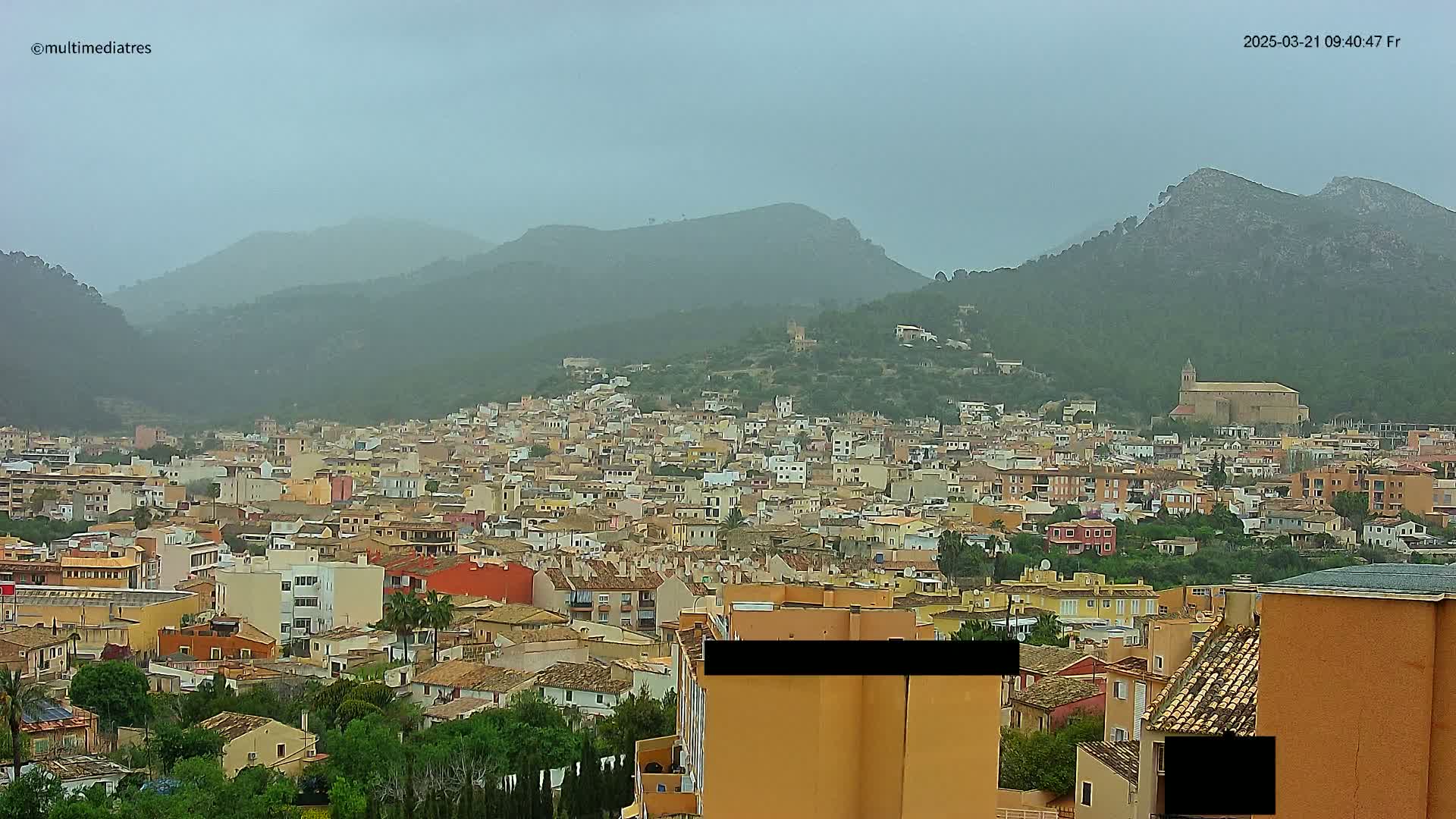 A sprawling town of low-rise buildings nestled in a valley between verdant hills under an overcast sky.