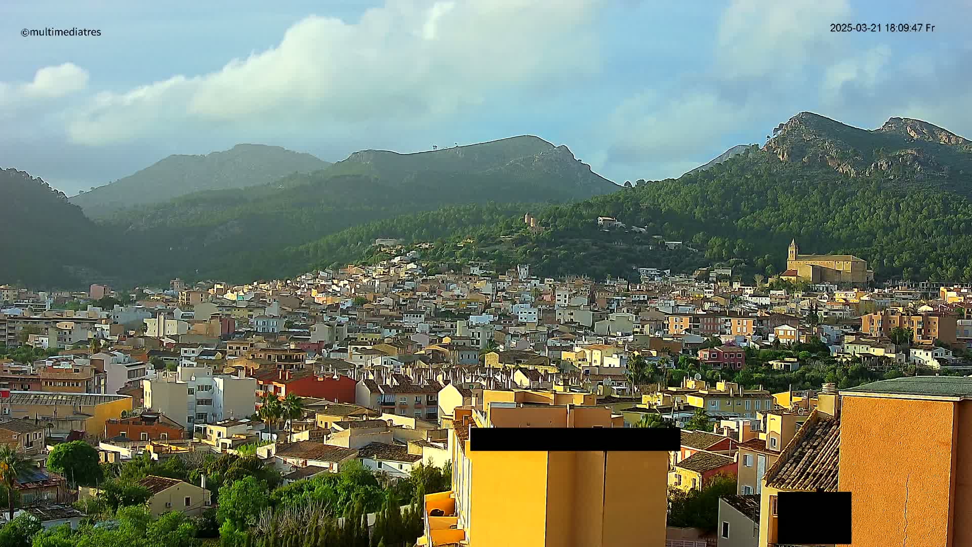 A sprawling town of low-rise buildings nestles at the foot of verdant, mountainous terrain under a partly cloudy sky.